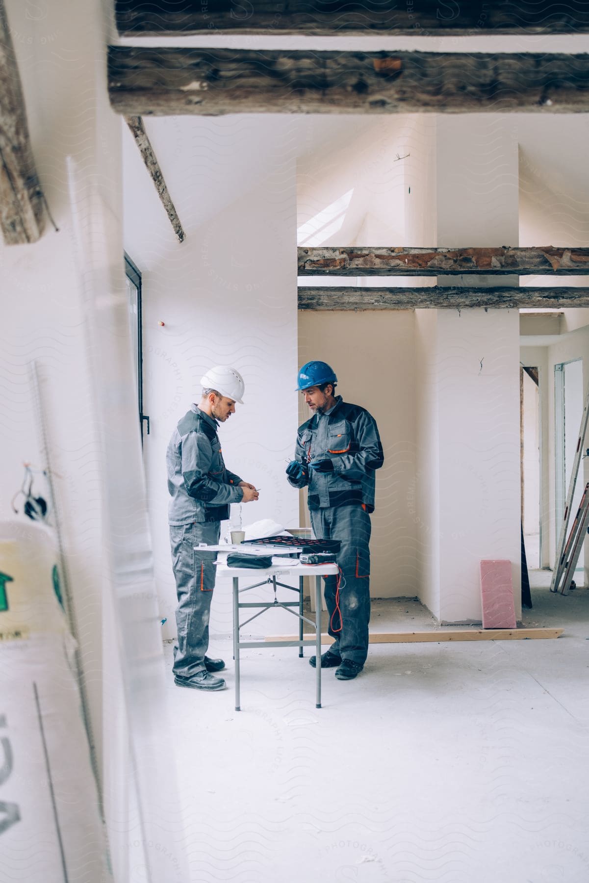 Two construction workers in work gear stand amidst renovation clutter (table and ladders) in a house under repair.