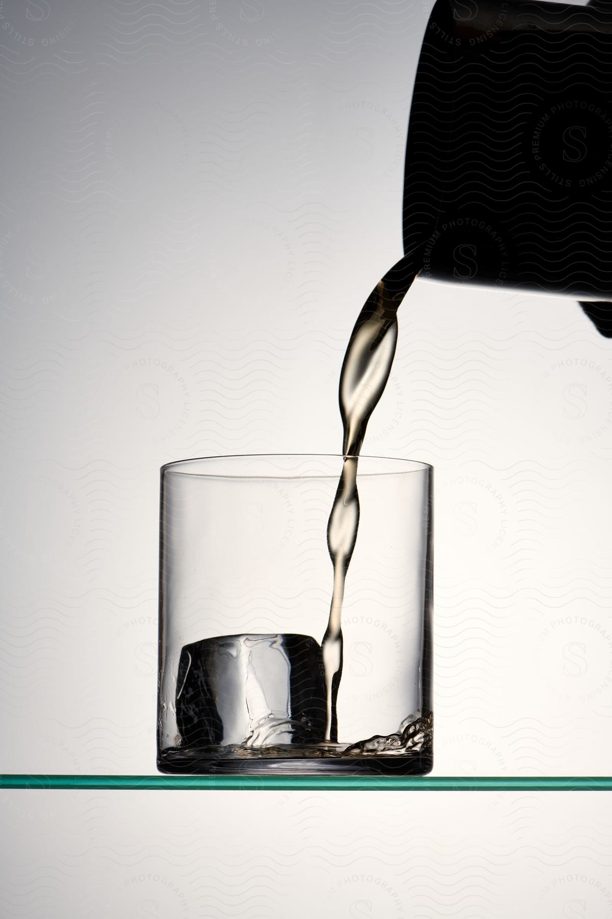 A beverage being poured into a water glass with an ice cube inside.