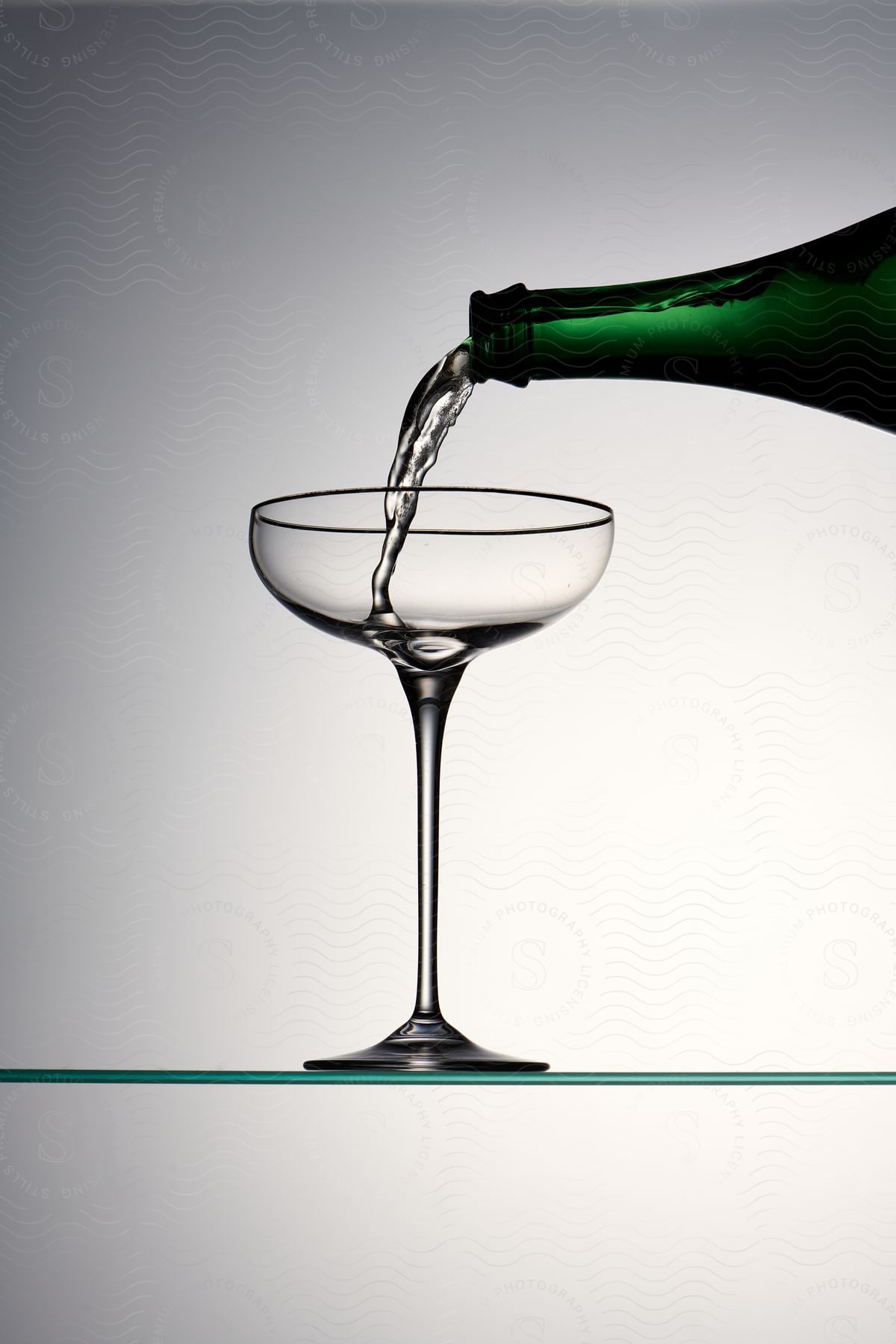 Liquid being poured from a green bottle into a martini glass on a clear surface with a gradient background.