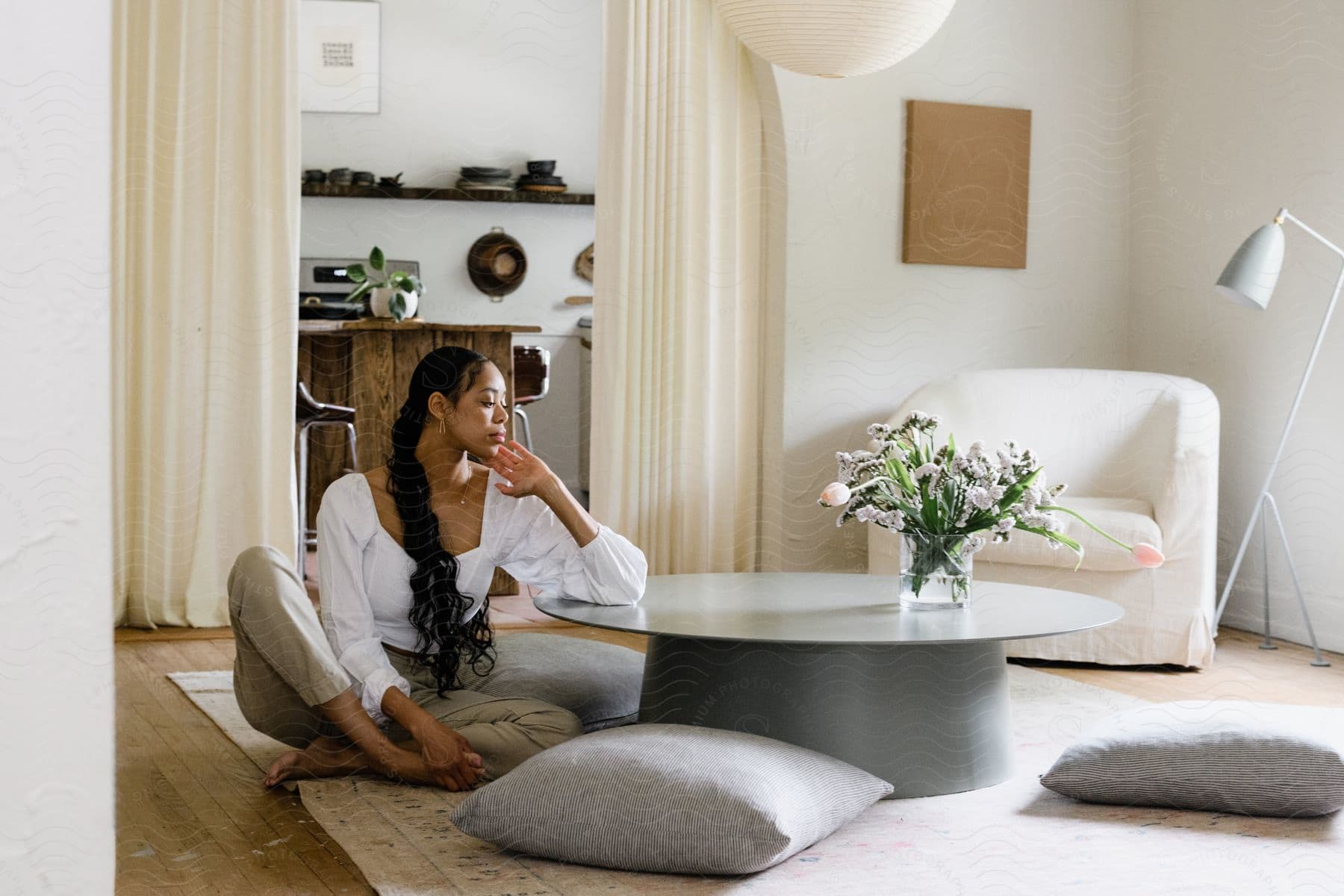 A woman with a white blouse and brown pants sits on the ground next to a coffee table in the living room.