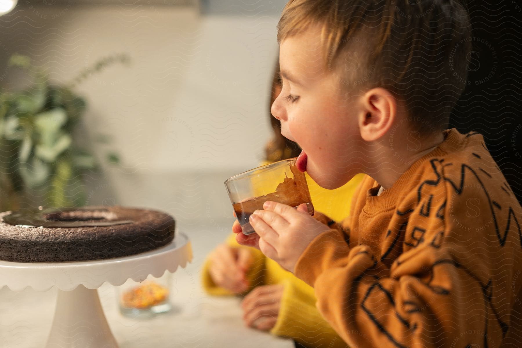 Stock photo of a boy in an orange shirt is licking the rim of a glass, with a chocolate cake on a stand in front of him.