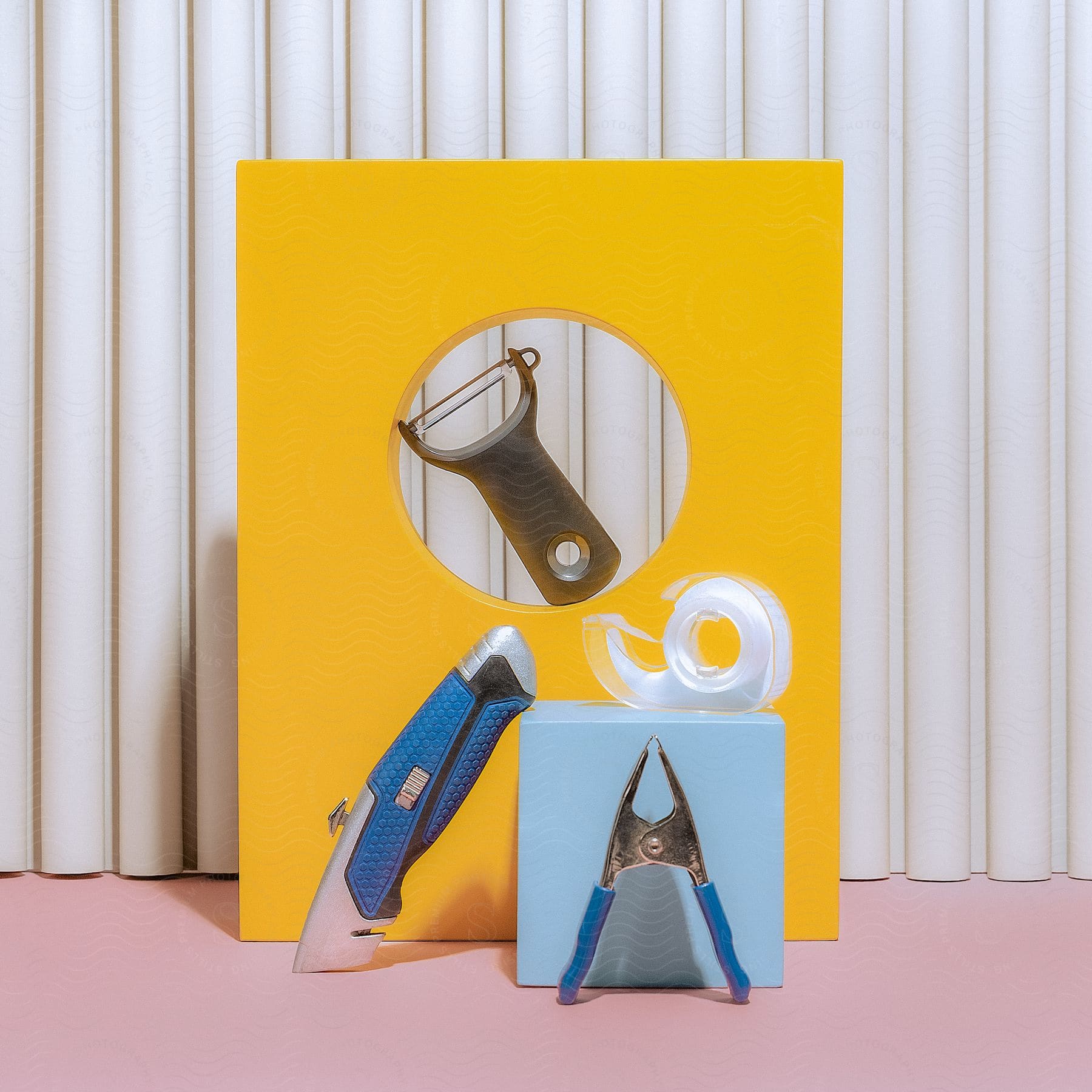 Office items creatively staged for a photo: a teal square with a box cutter, tape roll, blue-handled clamp; behind, a yellow rectangle with a hole, containing a potato peeler, against a pink table with a white ribbon background.