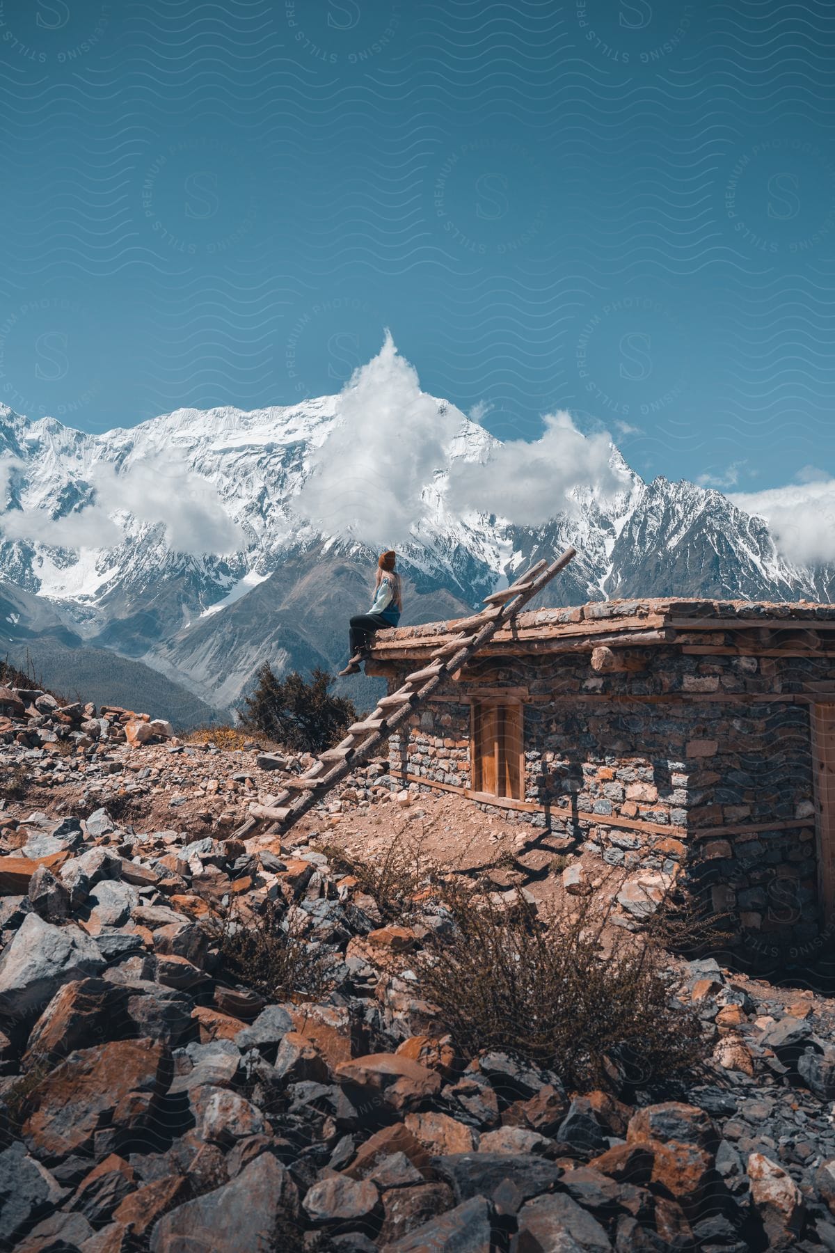 A woman sitting on the roof of a stone building with a backdrop of snowy mountain peaks and clouds.