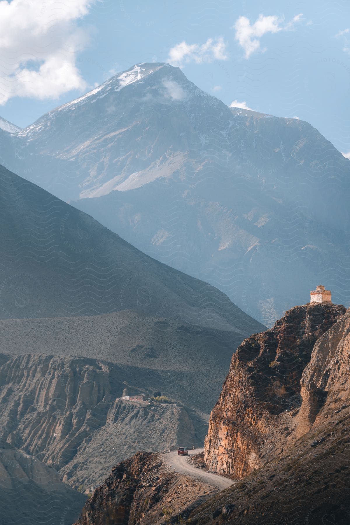 A truck travels around a curve on a mountain road with a small building on the mountaintop and mountains in the distance