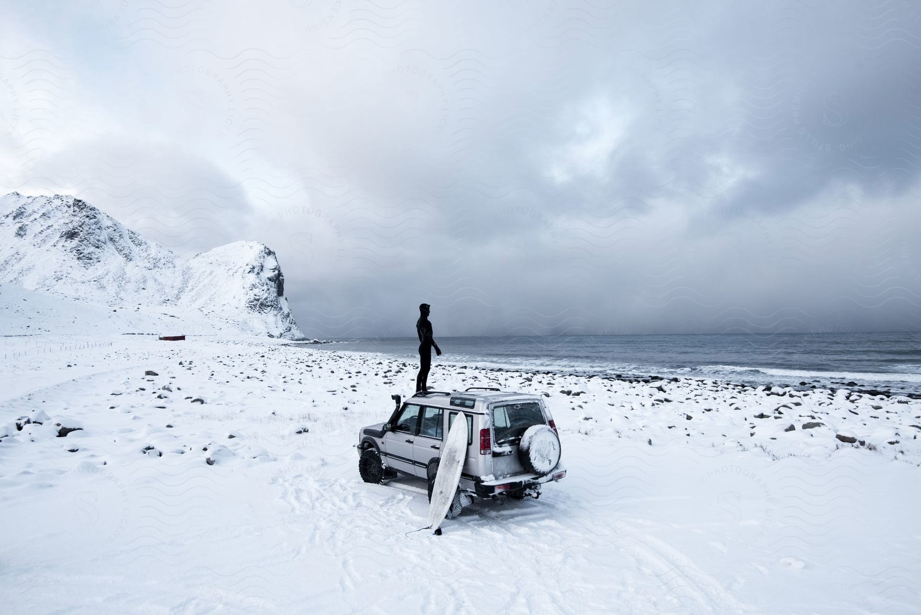 A man stands on his van with a surfboard leaning against the side parked on a snow covered beach as he looks at the water