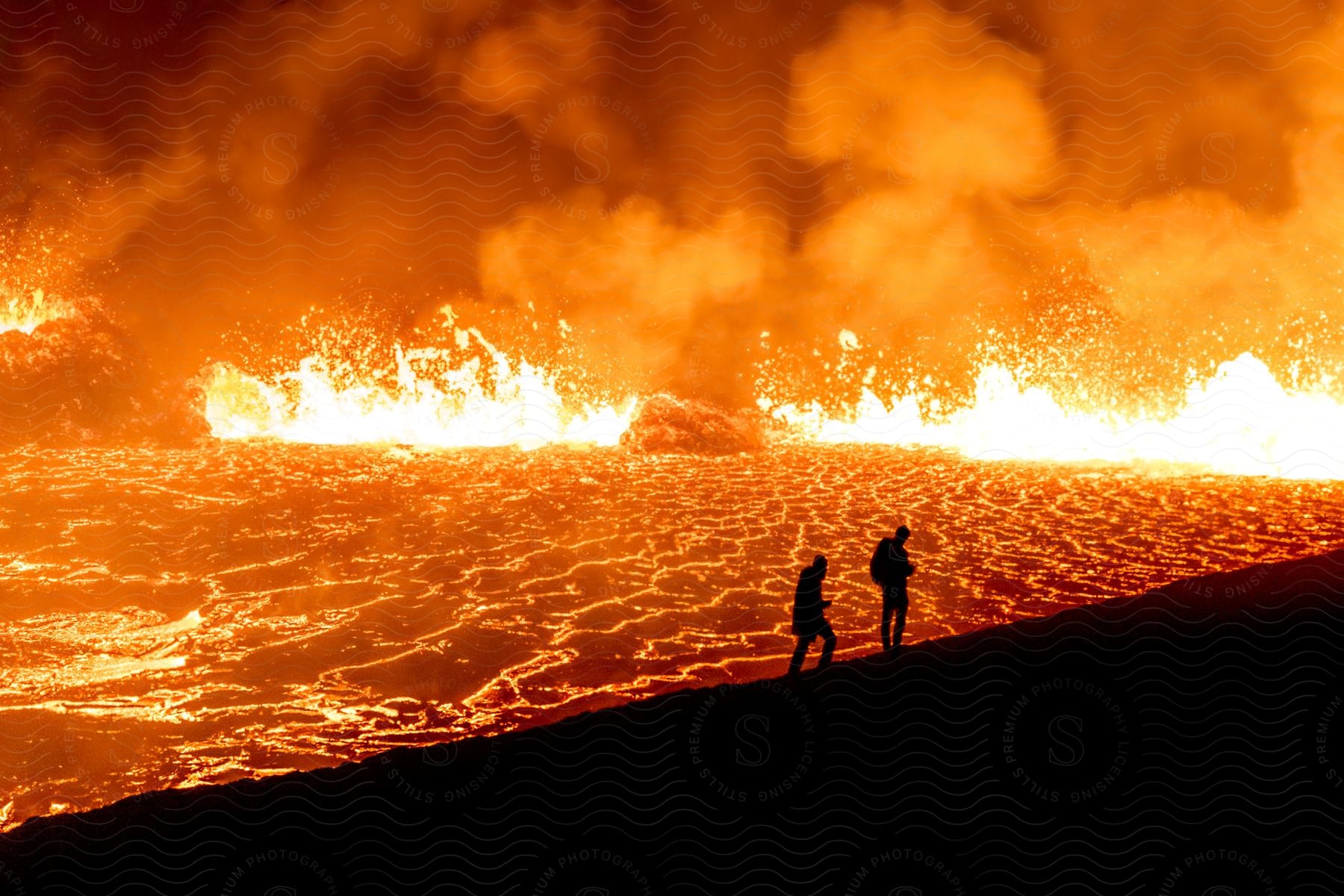 Silhouettes of two people standing on a hill overlooking glowing splashing lava in an erupting volcano