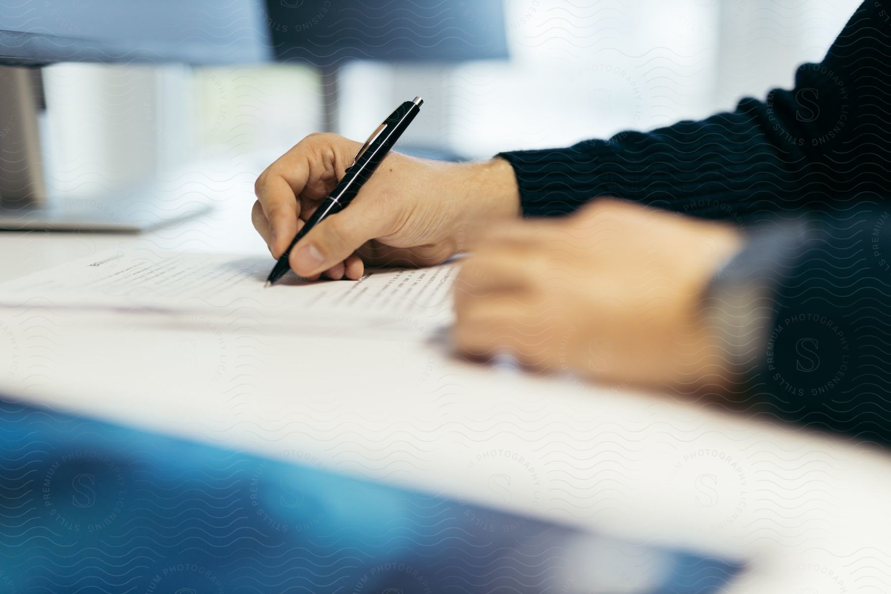 close-up of a person's hands in a dark sweater writing on paper with a black pen, indoors.