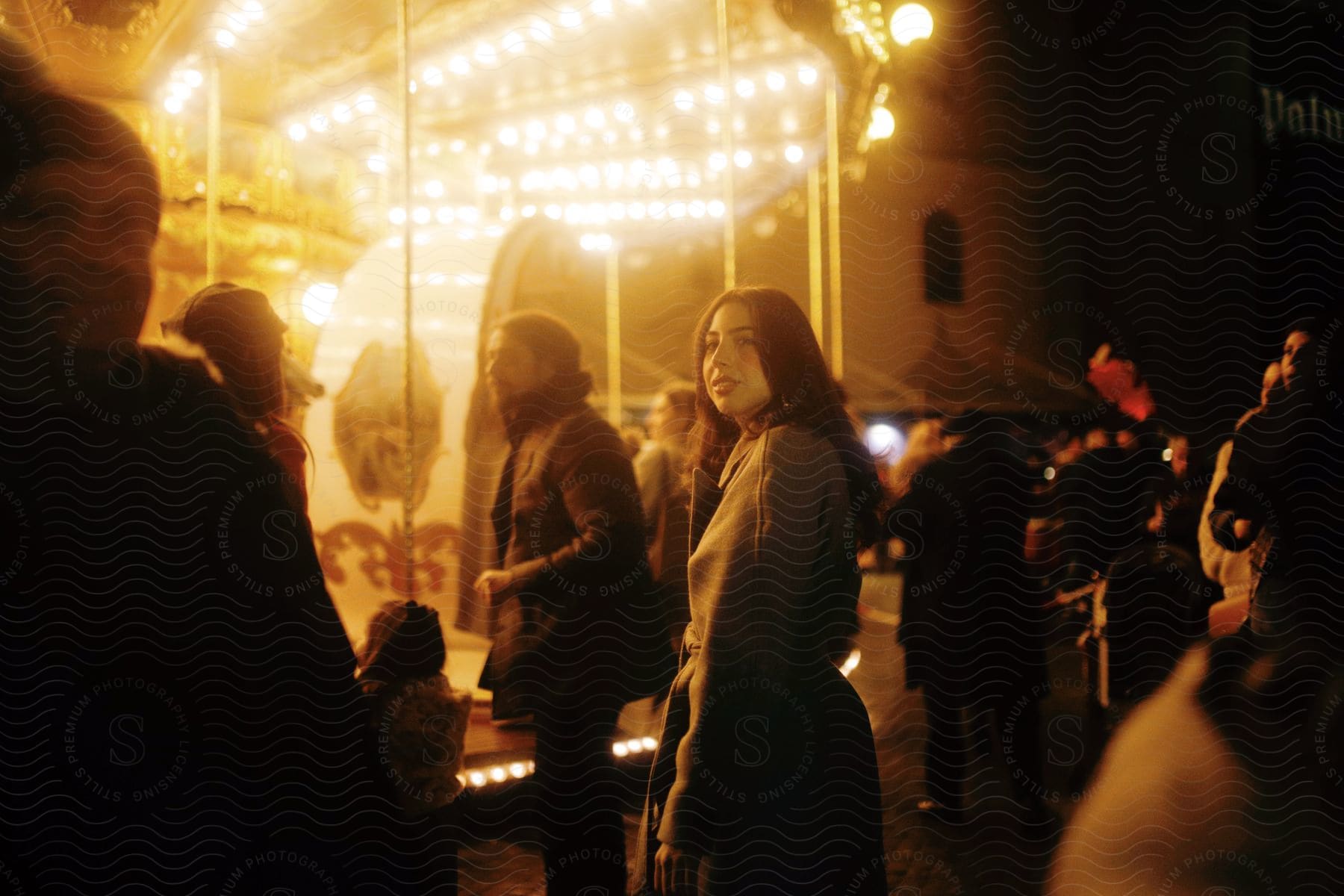 A girl is standing in front of a carousel at night.