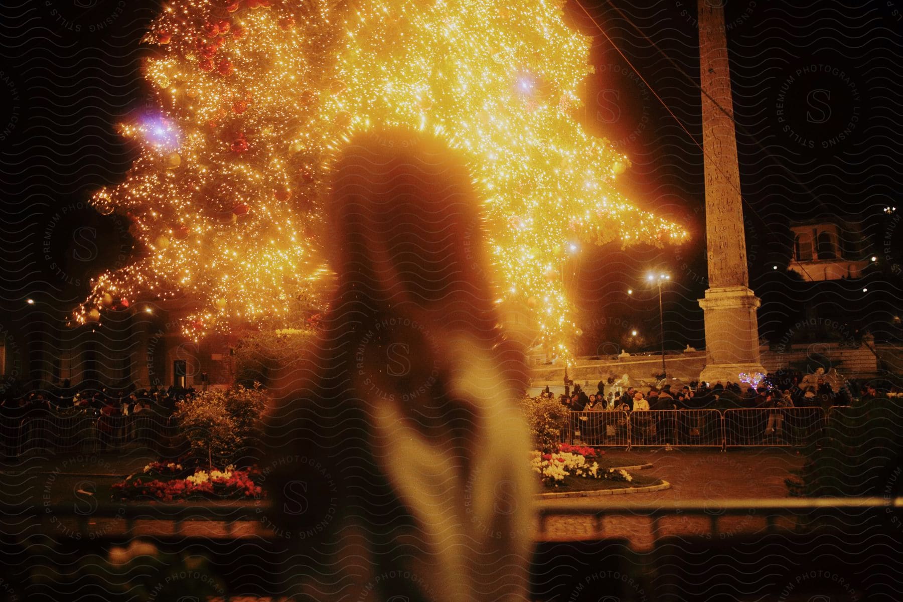 A woman watches from a balcony as people are standing in a park at night watching the glowing fireworks explode across the darkness