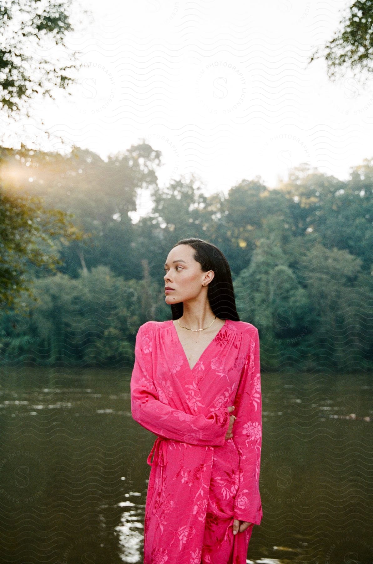 A woman wearing a pink dress is standing along the coast of a river in the woods with her head turned to the side