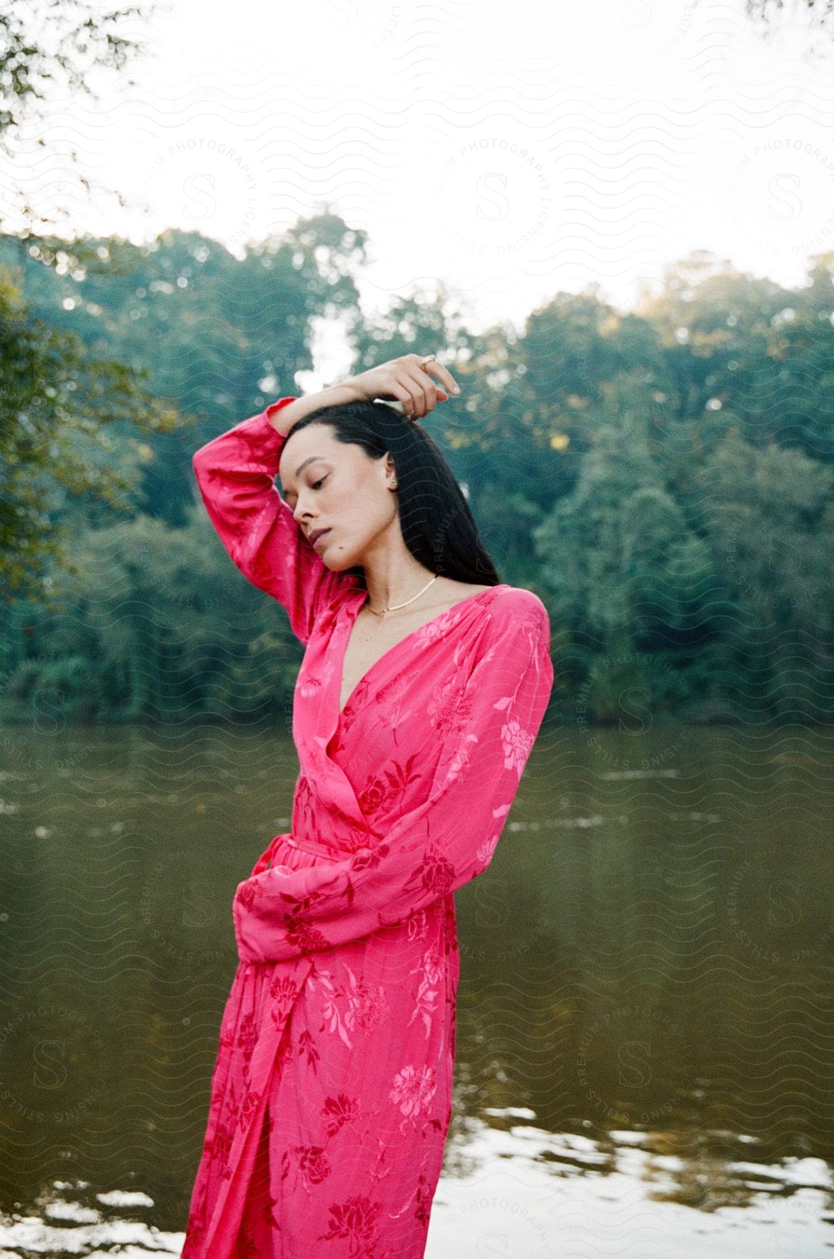 A woman wearing a pink dress is standing along the coast of a river in the woods with one arm on her head turned to the side