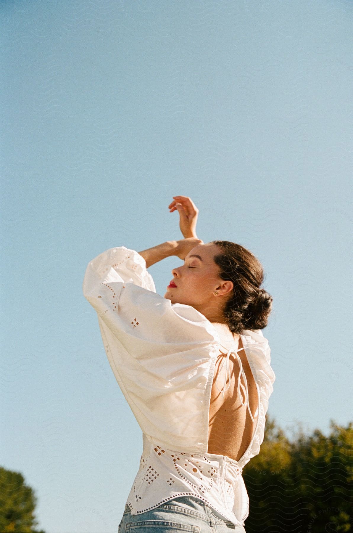 A young woman models a white, backless shirt while standing outside on a sunny, summer day.