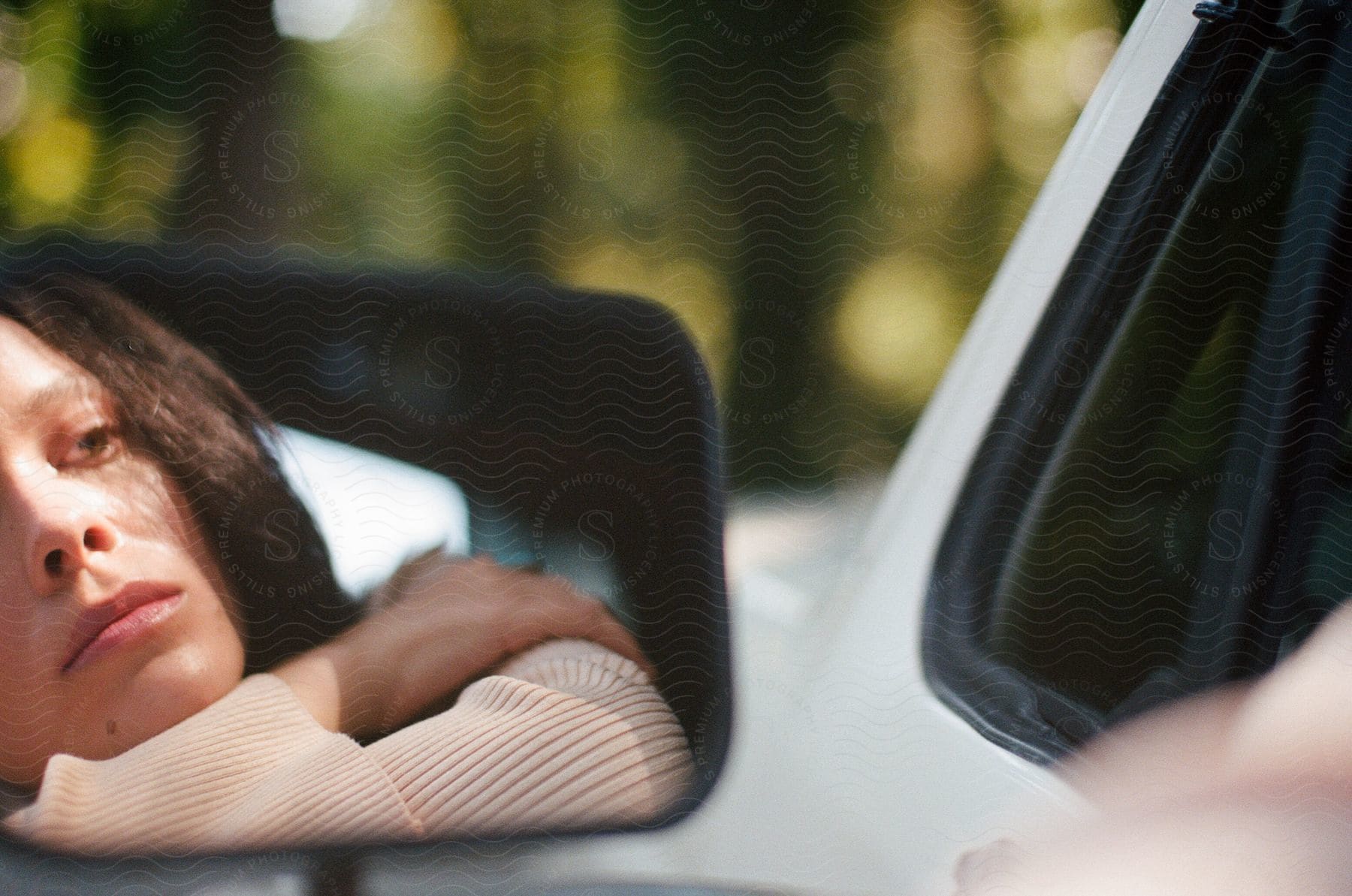 A young woman in the driver's seat of a parked car, leaning off the window with her arms crossed while reflected in the mirror.