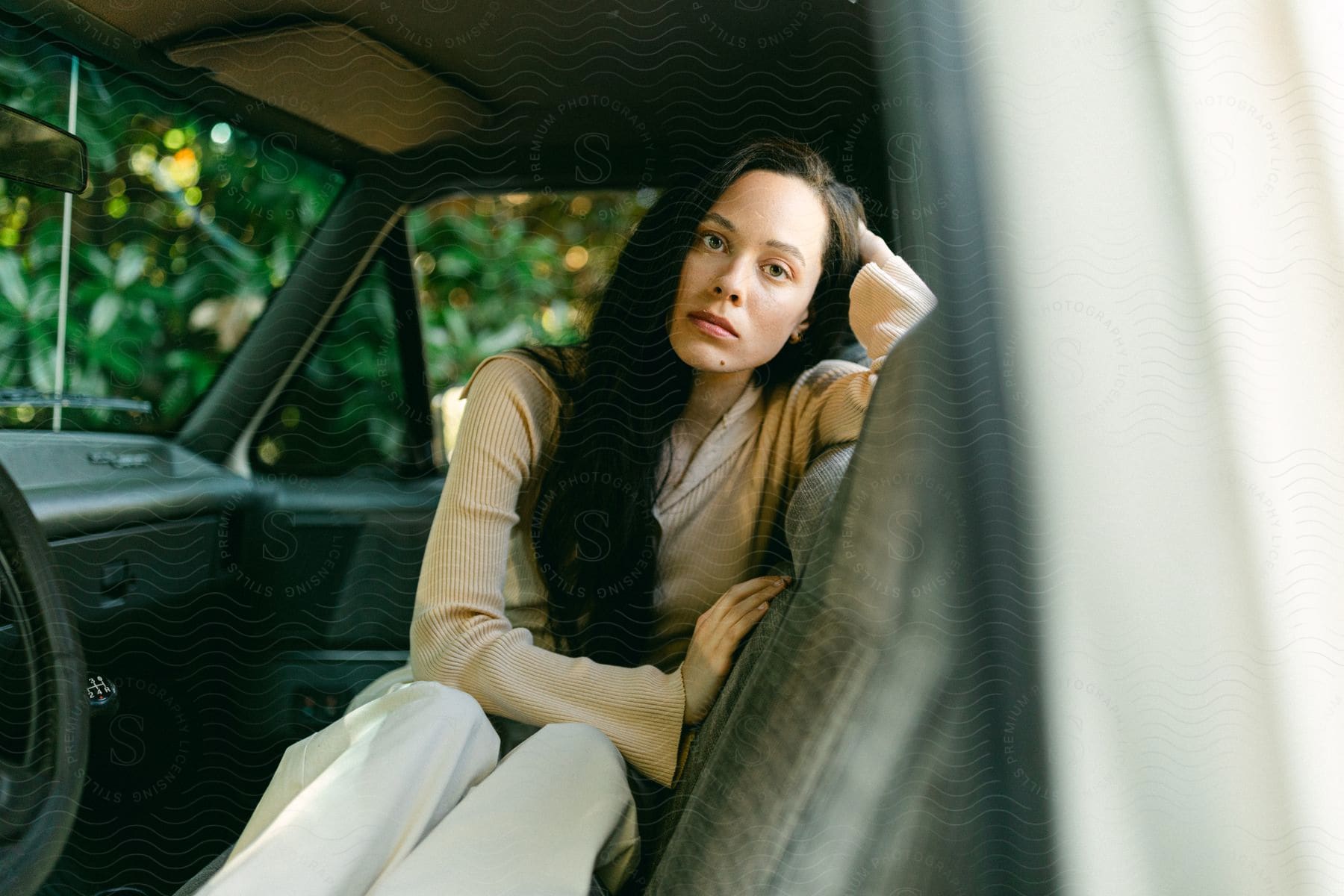 Woman leans against the side of a bench seat facing the driver's side door in the front of an old green vehicle in front of a large bush.