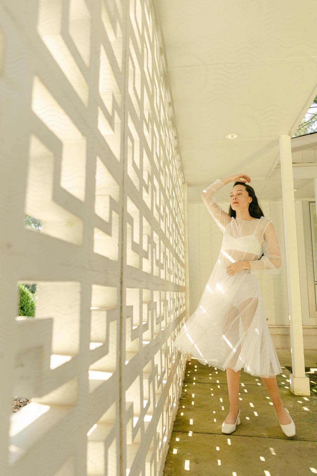 A woman wearing a sheer white gown stands near a wall as she holds one arm over her head