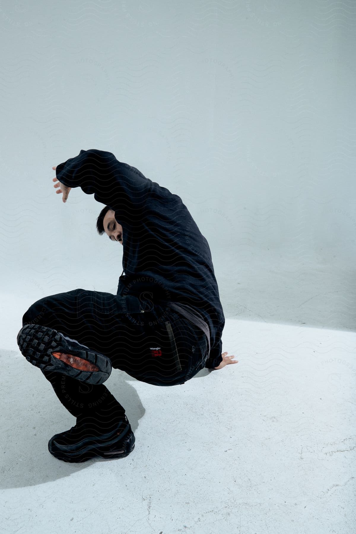 A person in black clothes is performing a dynamic dance move on a light grey floor against a grey background.