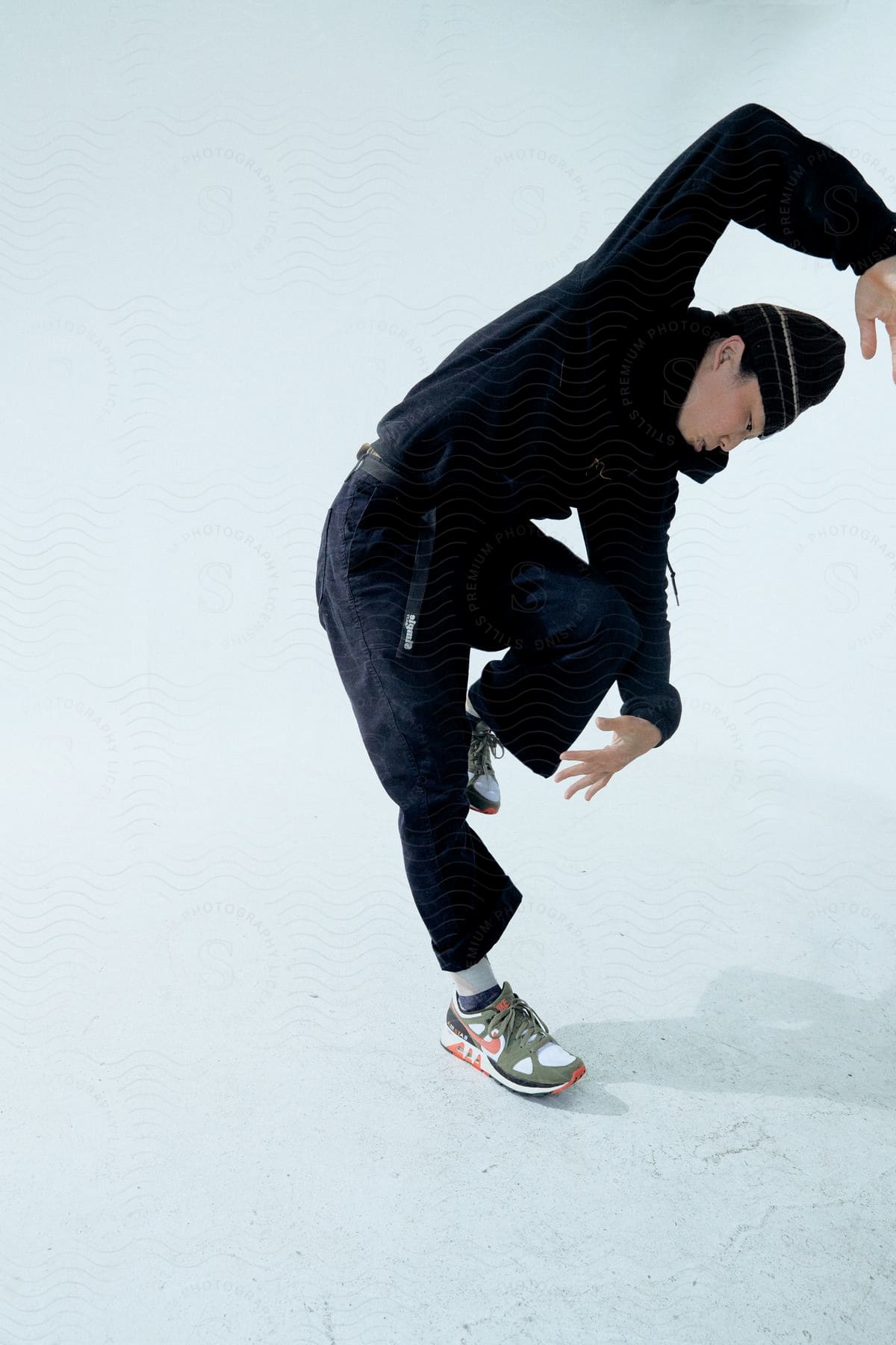 A boy in all black clothing posing with his leg bent and his arms raised on an all white background