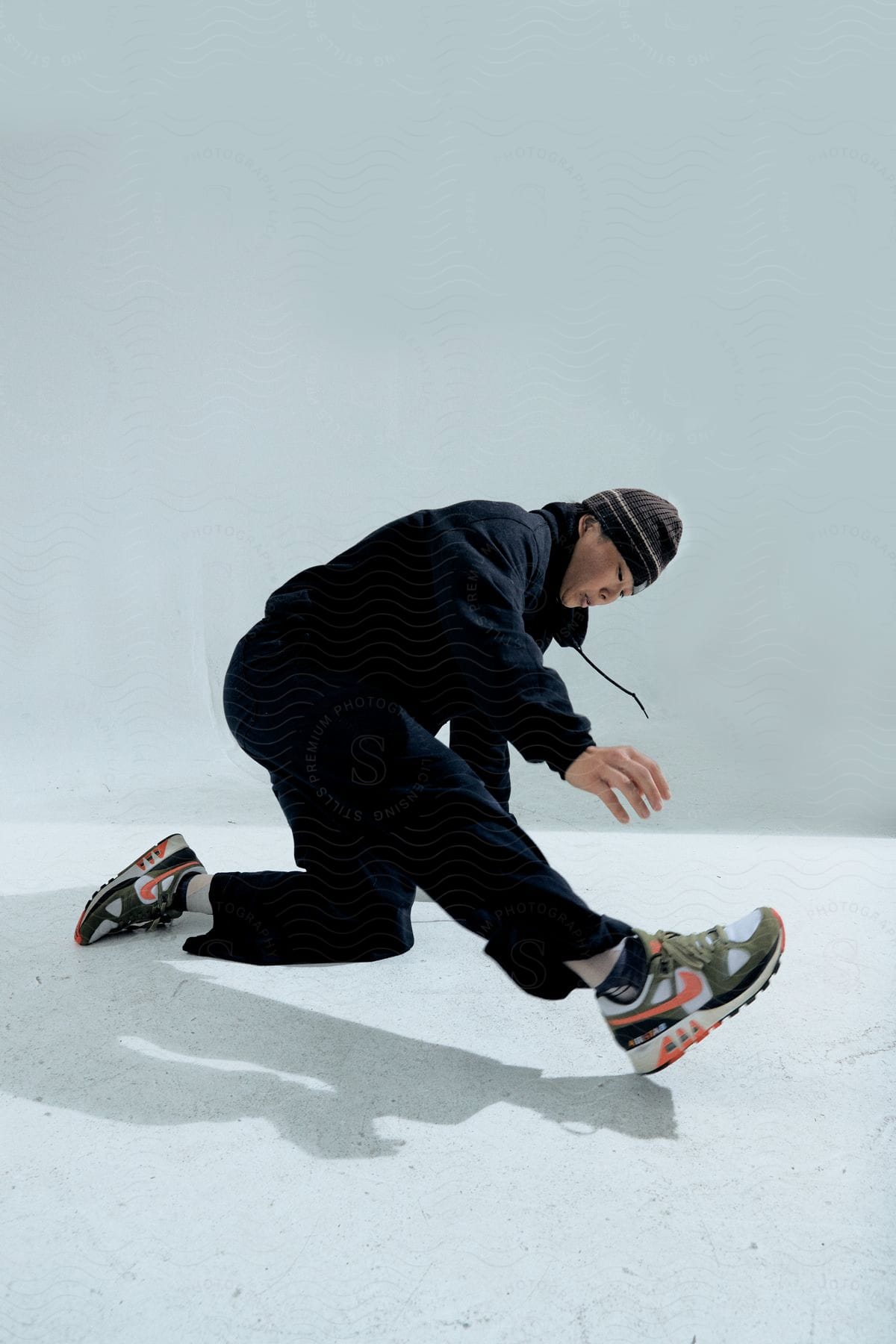 Man wearing black pants, black hoodie and black beanie crouches on white background