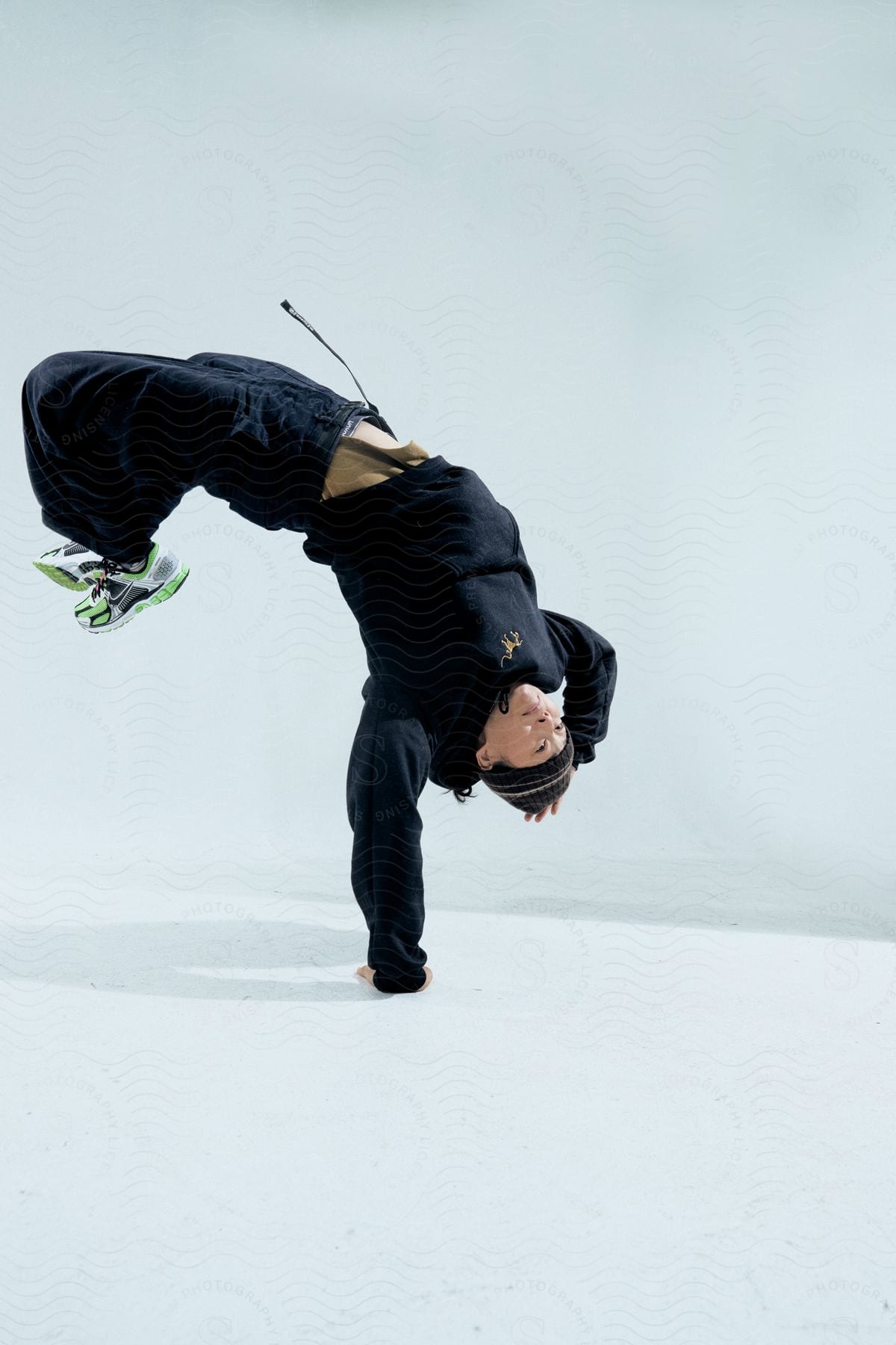 Stock photo of woman performing an acrobatic movement, with her legs raised in the air and dressed in black clothing and green and white sneakers, against a light background.