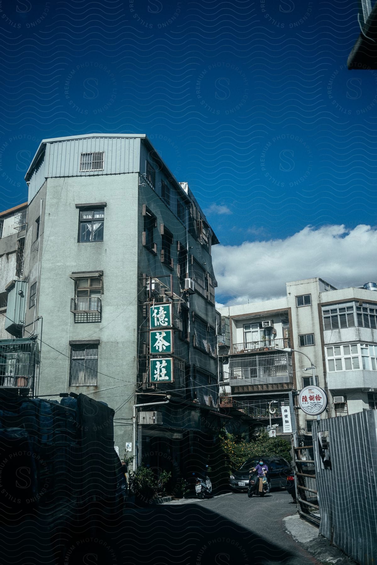 A person sits on a bike near cars on the street outside of apartment buildings