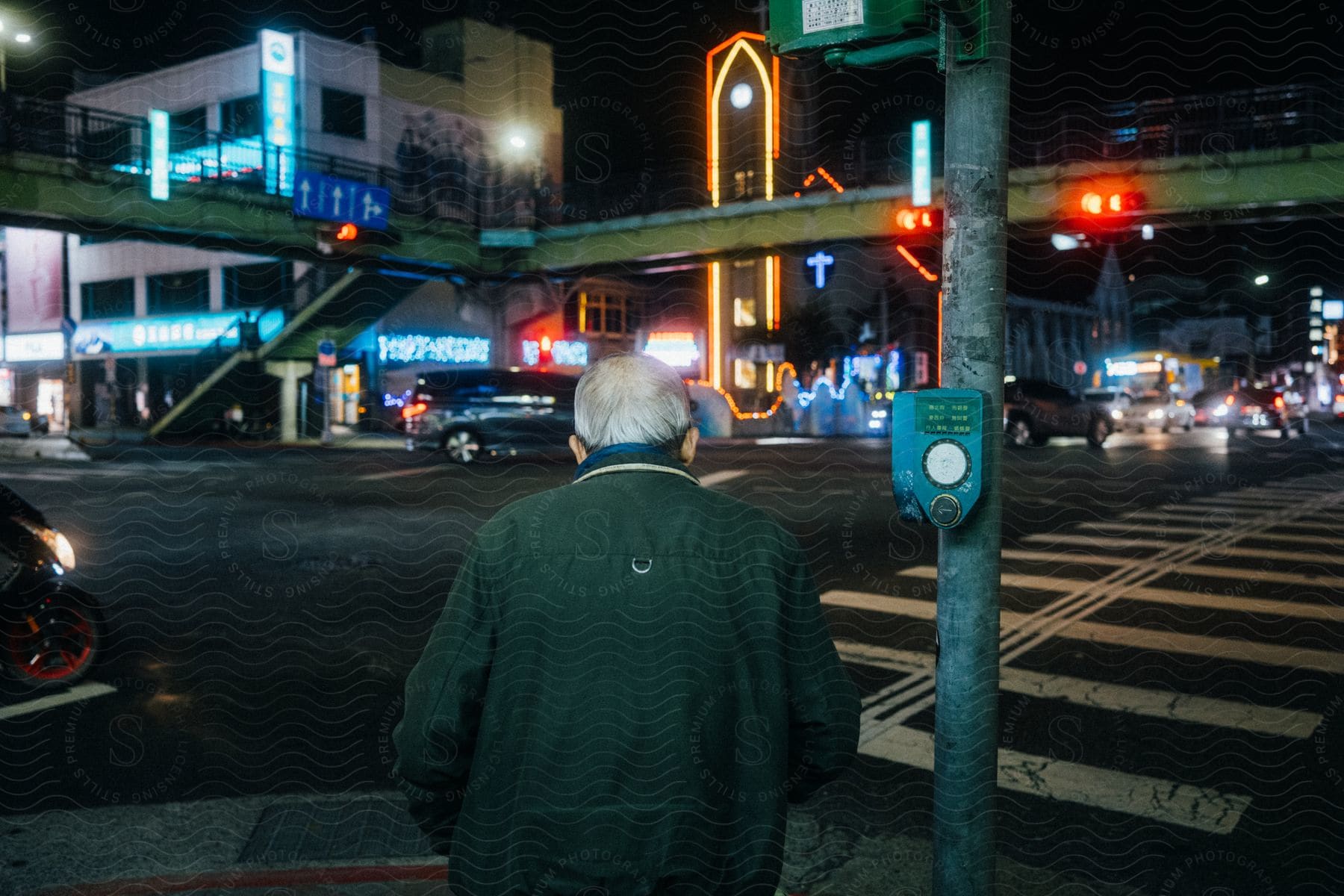 A man stands at the crosswalk of a downtown street as cars pass and neon signs glow in the darkness