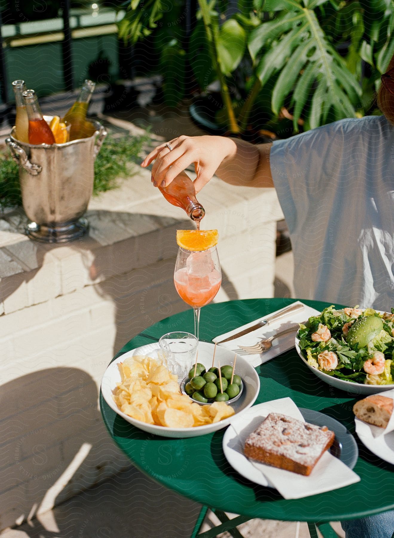 A woman sits at an outdoor table with a salad and plates of food as she pours a drink into a glass from a bottle