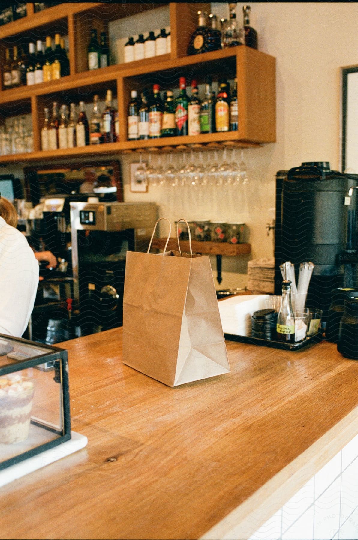 A brown bag is on the counter as an employees hands are seen working below a shelf filled with bottles of alcohol