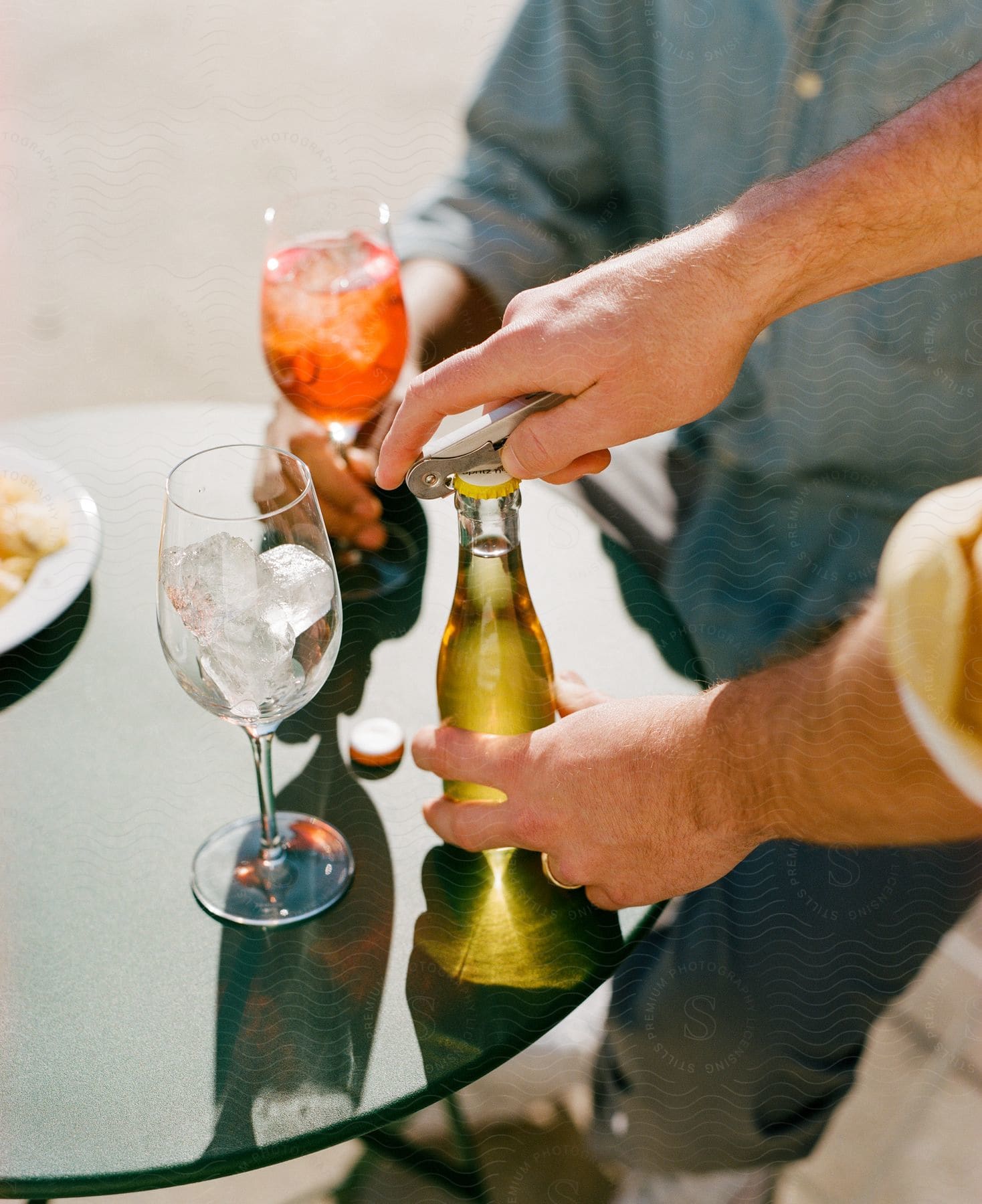 A man is standing at a table opening a bottle to pour into a glass of ice