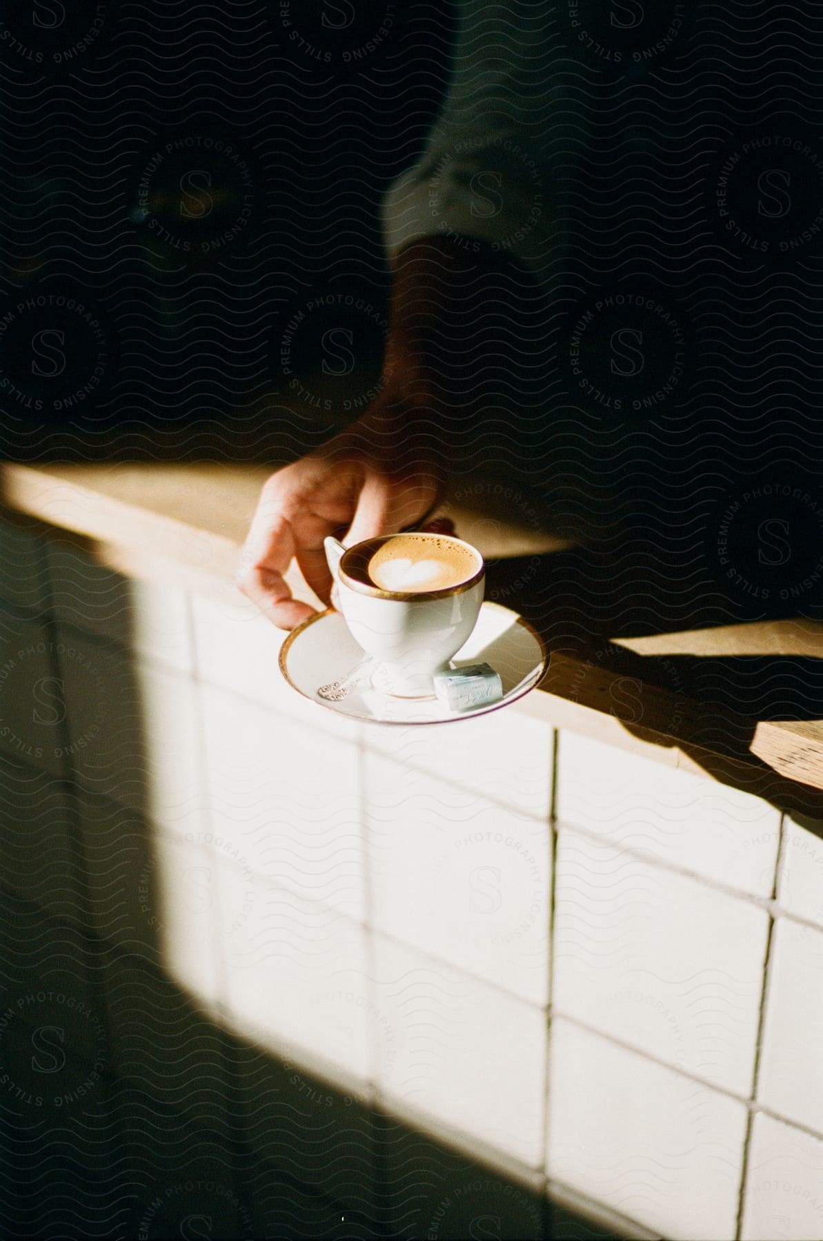 A persons hand is holding a cup of coffee on a saucer over the edge of a counter