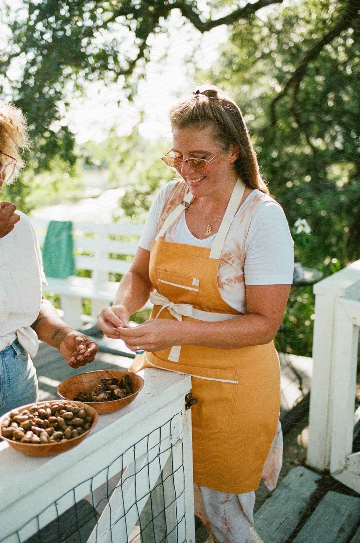 Woman in an orange apron shelling nuts on a porch with greenery in the background.