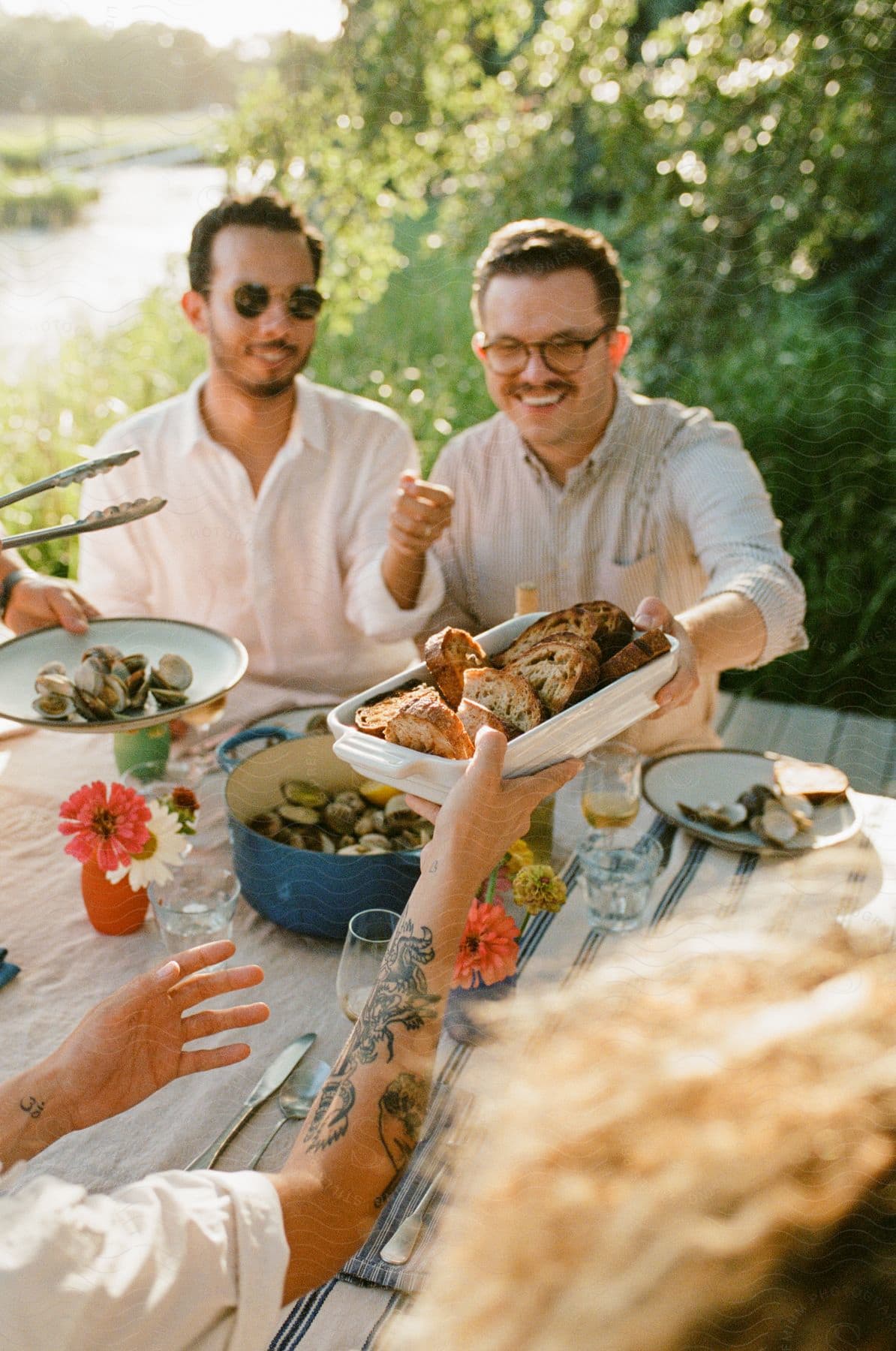 A group of male friends smiling and sharing a pot of steamers and garlic bread at a riverside table