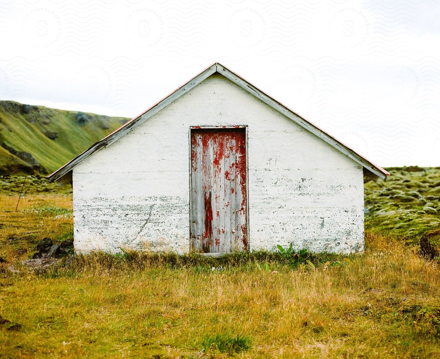 An old white building with a door sits in a field with a grass covered slope in the distance