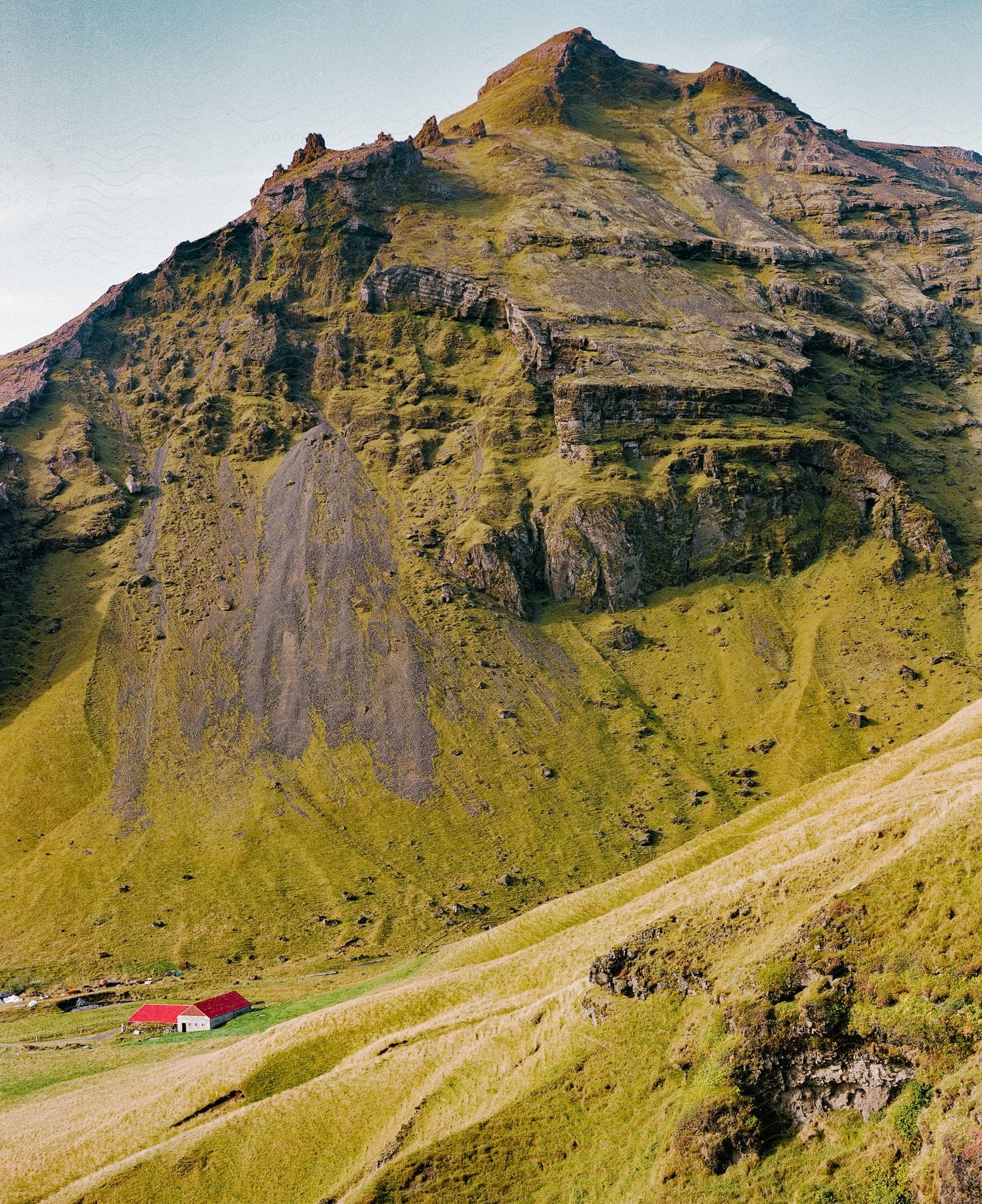 A farmhouse sits at the base of a steep rocky mountain on a sunny day.