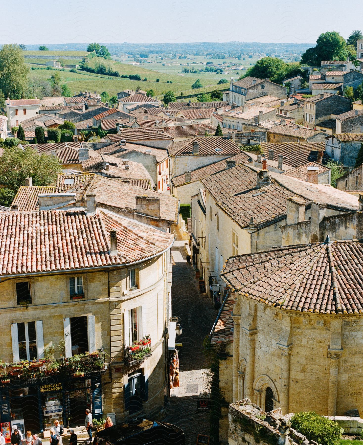 People stand in front of a store in Saint Emilion France among buildings on a narrow street and grass covered hills in the distance