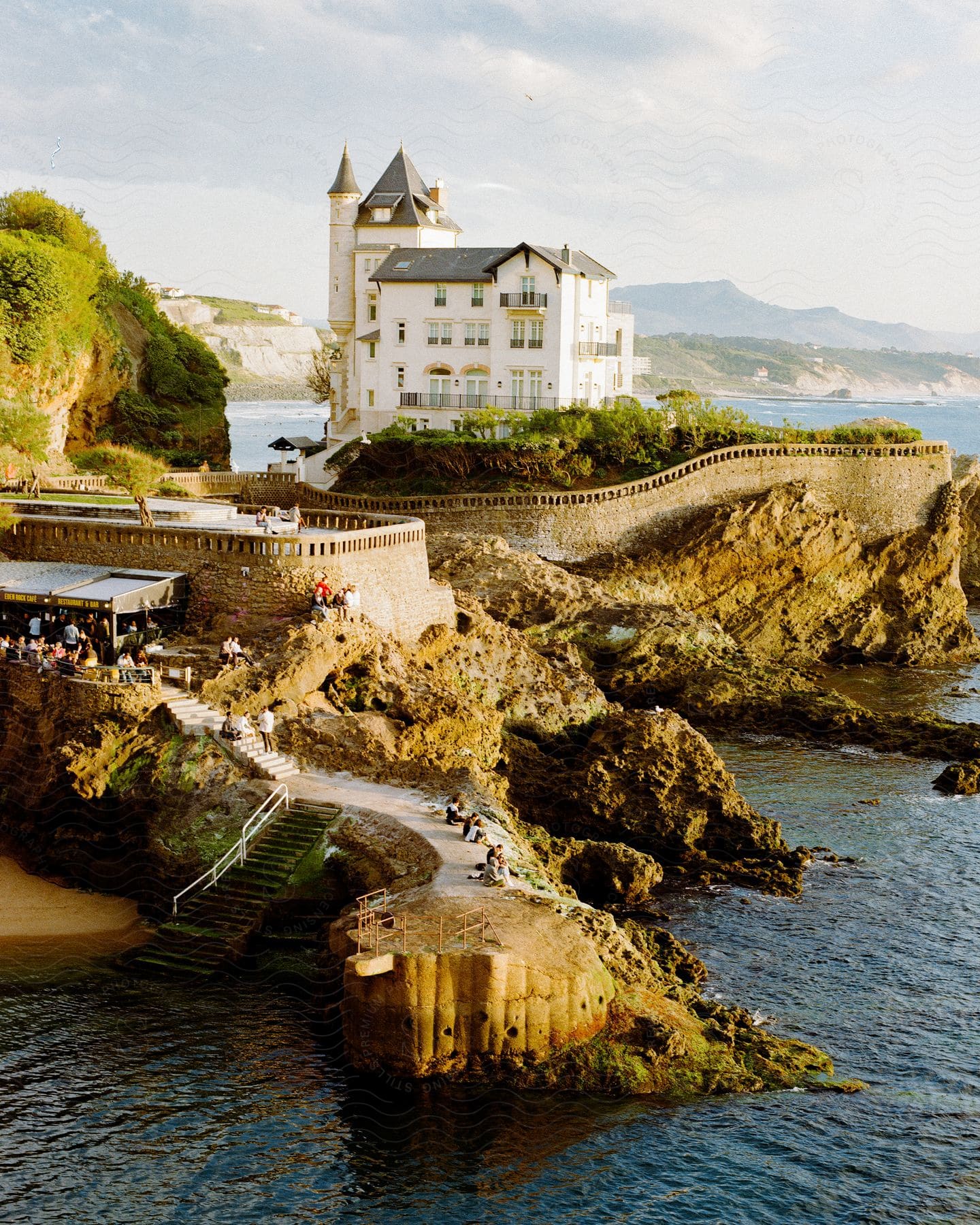 People are outside at a French chateau on the coast as waves splash against rocks