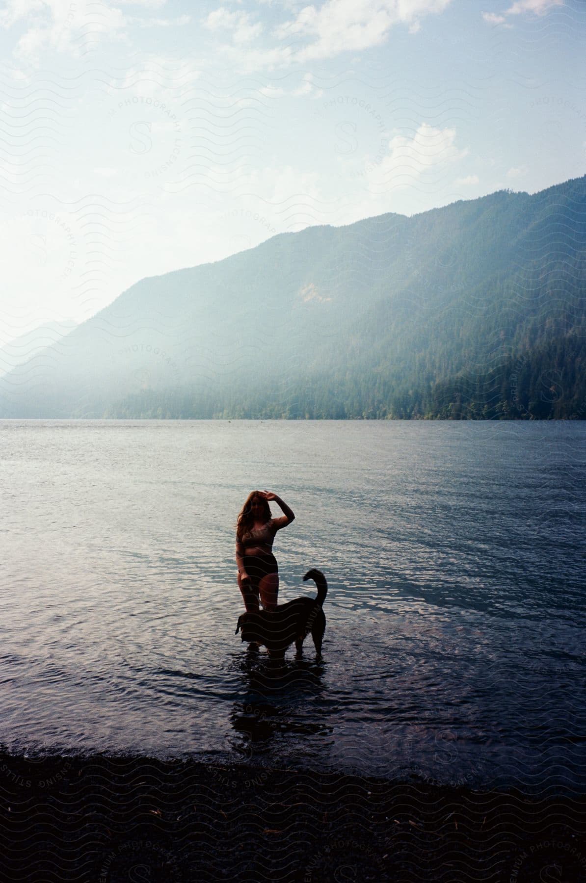 A woman wades in a mountain lake with her dog on a partly sunny summer day.