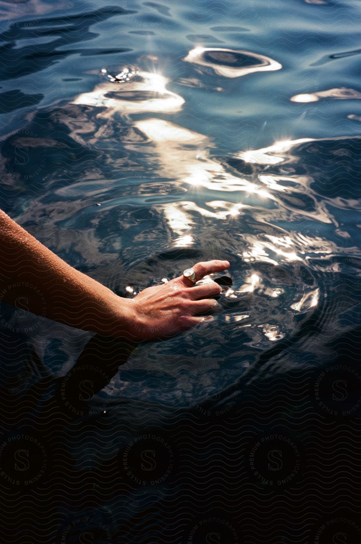 A woman's hand with a silver ring on her finger reaches into the water as sunlight reflects on the ripples and waves