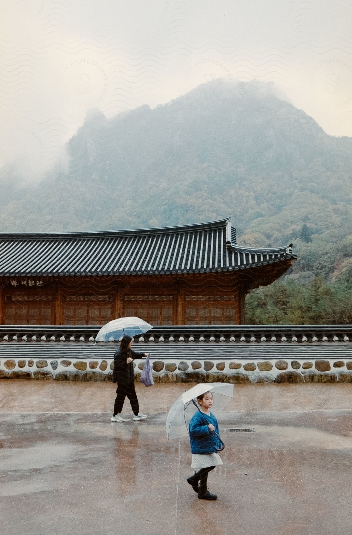 Mother & daughter walking with umbrellas in the rain by a building in front of a mountain