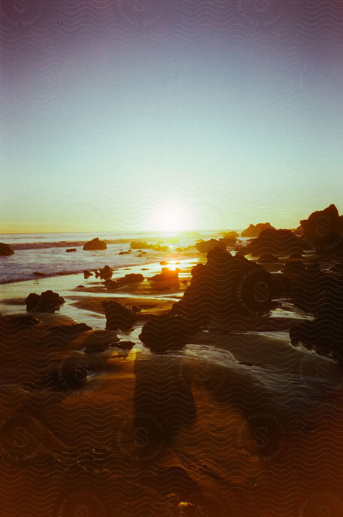 The sun is setting over an ocean beach scattered with craggy boulders.