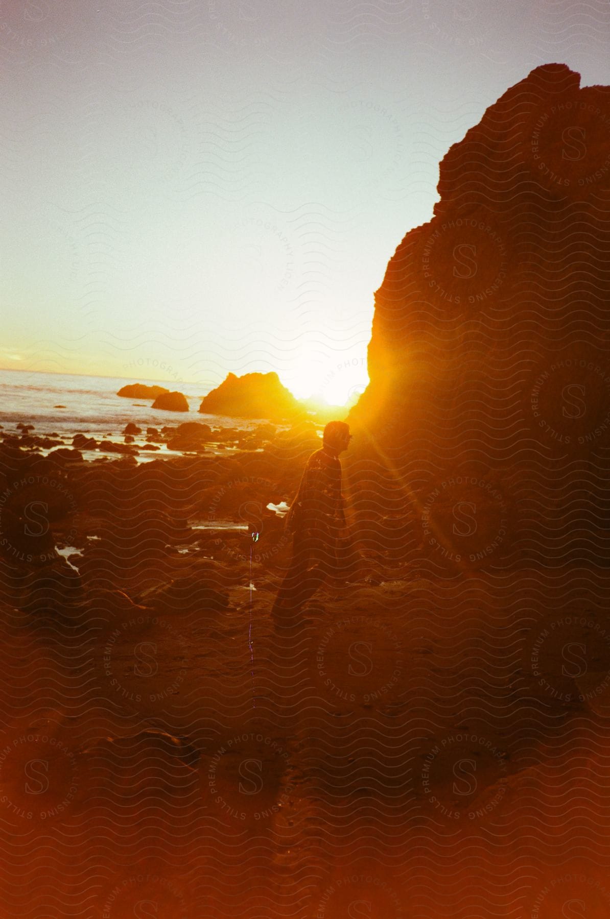 A man is walking near a promontory along the coast as the sun shines brightly in the distance with silhouettes of rocks in the water