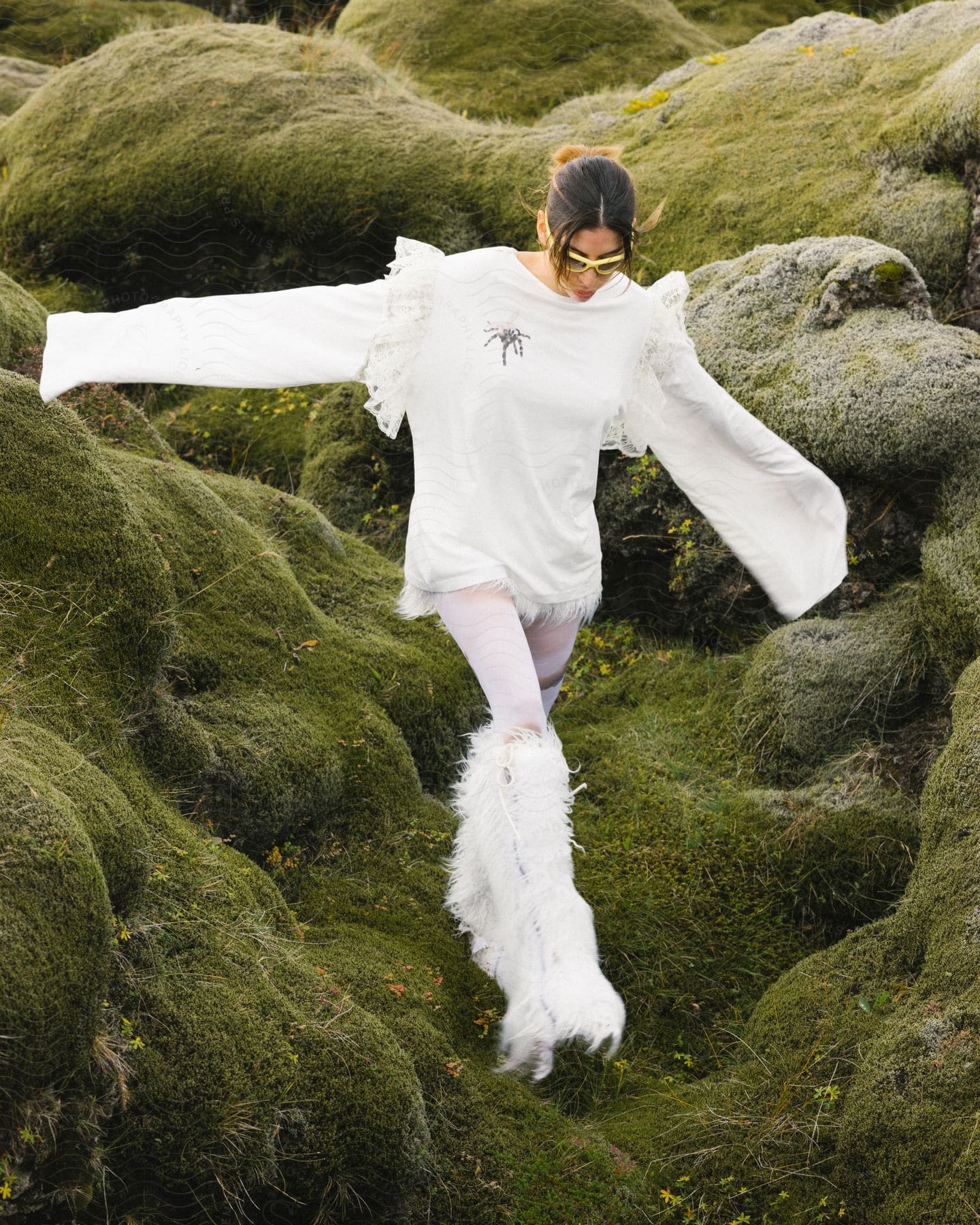 A woman wearing white knee high feathered boots and a long sleeve white tunic walking on green covered rocks