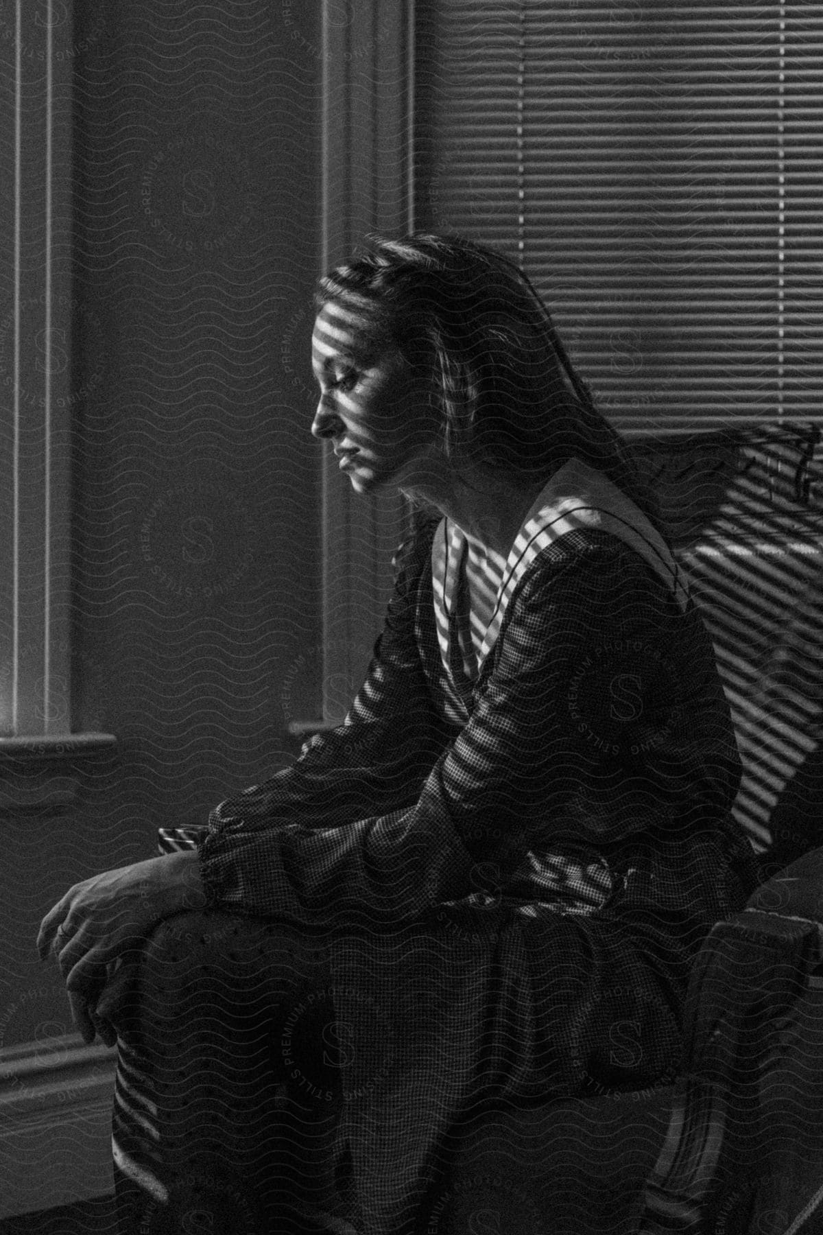 A woman is sitting in an armchair and lights from the window illuminate her face, which appears sad.