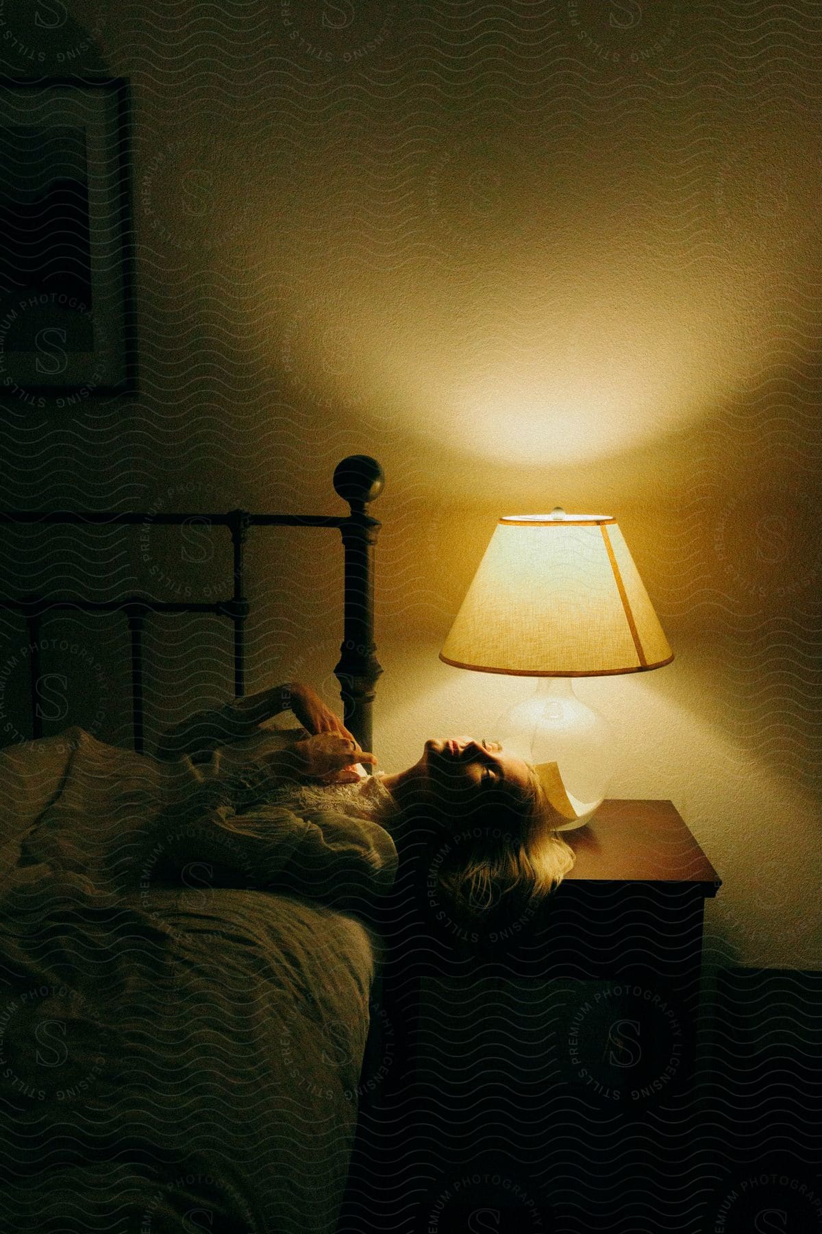 A woman lying across a bed with her head on the night table and hands held on her chest in an awkward manner