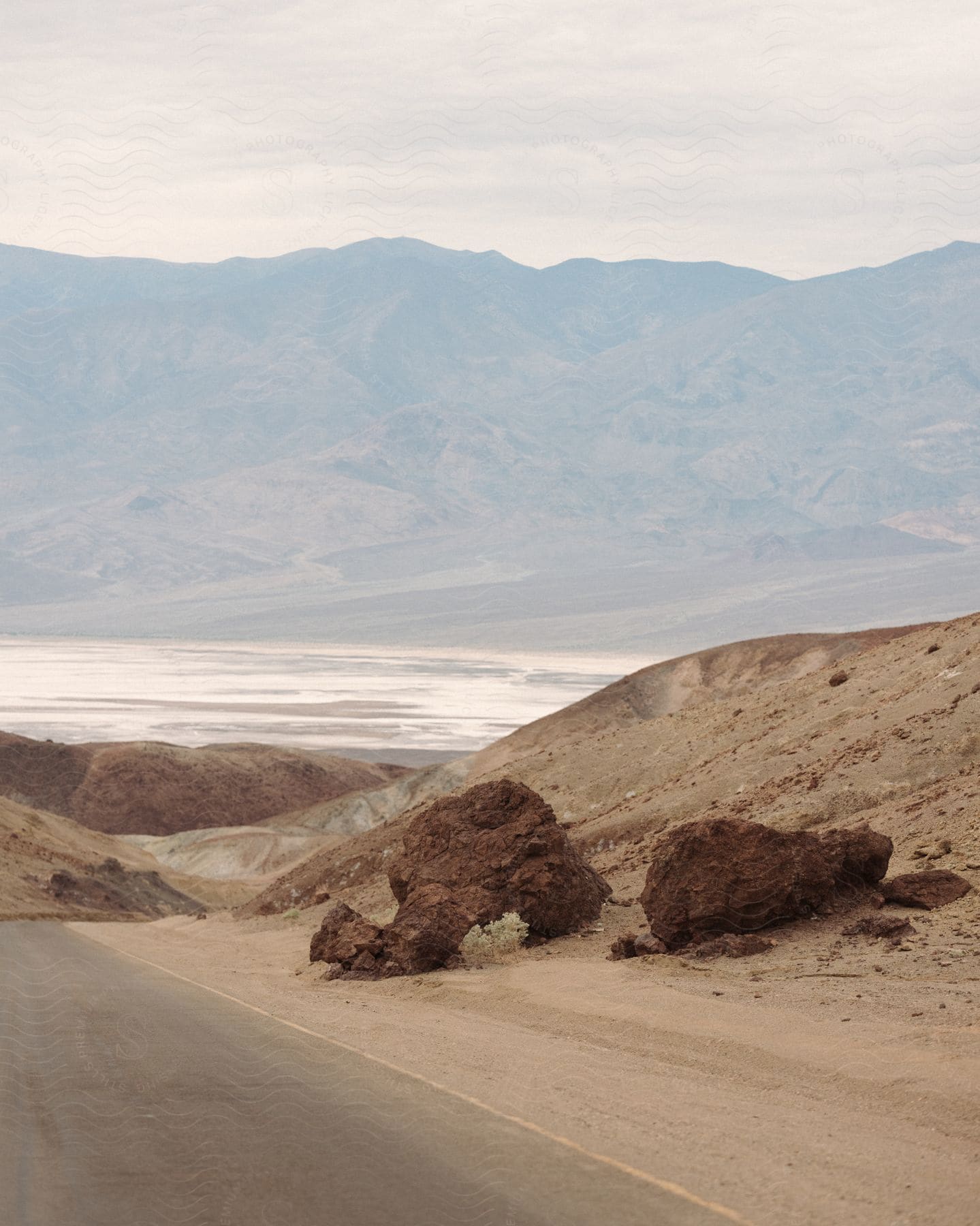 A region with an asphalt road next to rocks that are on a sandy ground and in the background large mountains