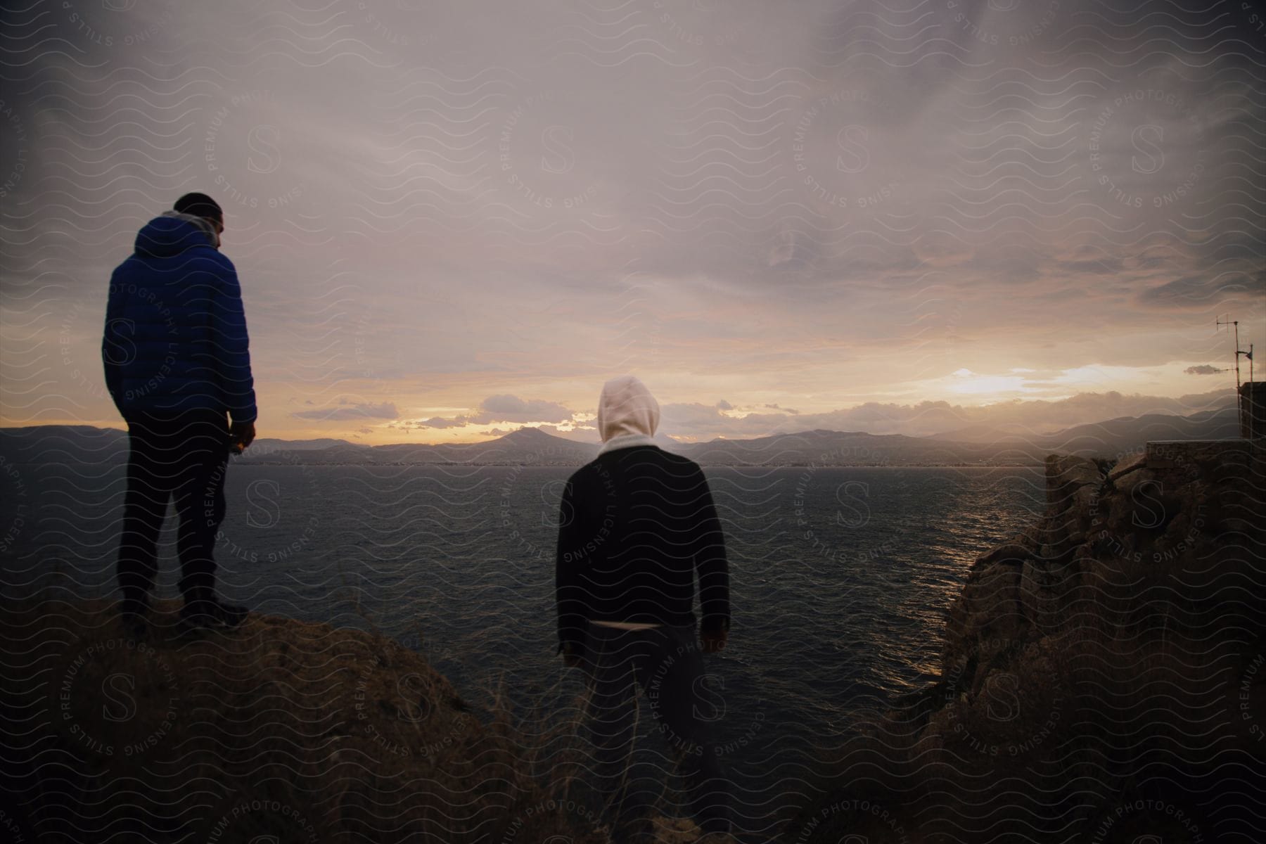 Two people looking off a cliff at the sunset in the distance on the ocean.