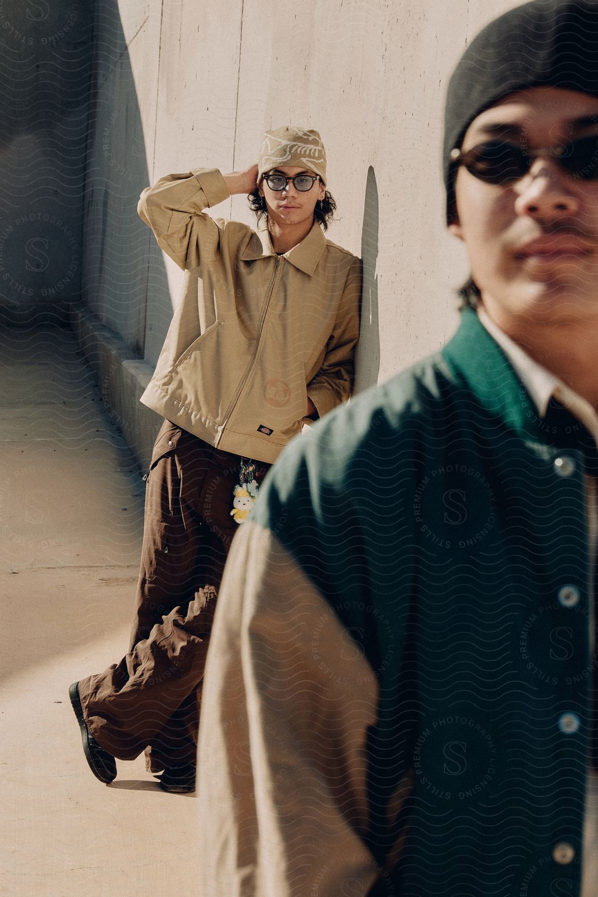 Two man in casual clothing posing against a concrete structure, one in foreground with a black beanie and the other in background adjusting a tan beanie.