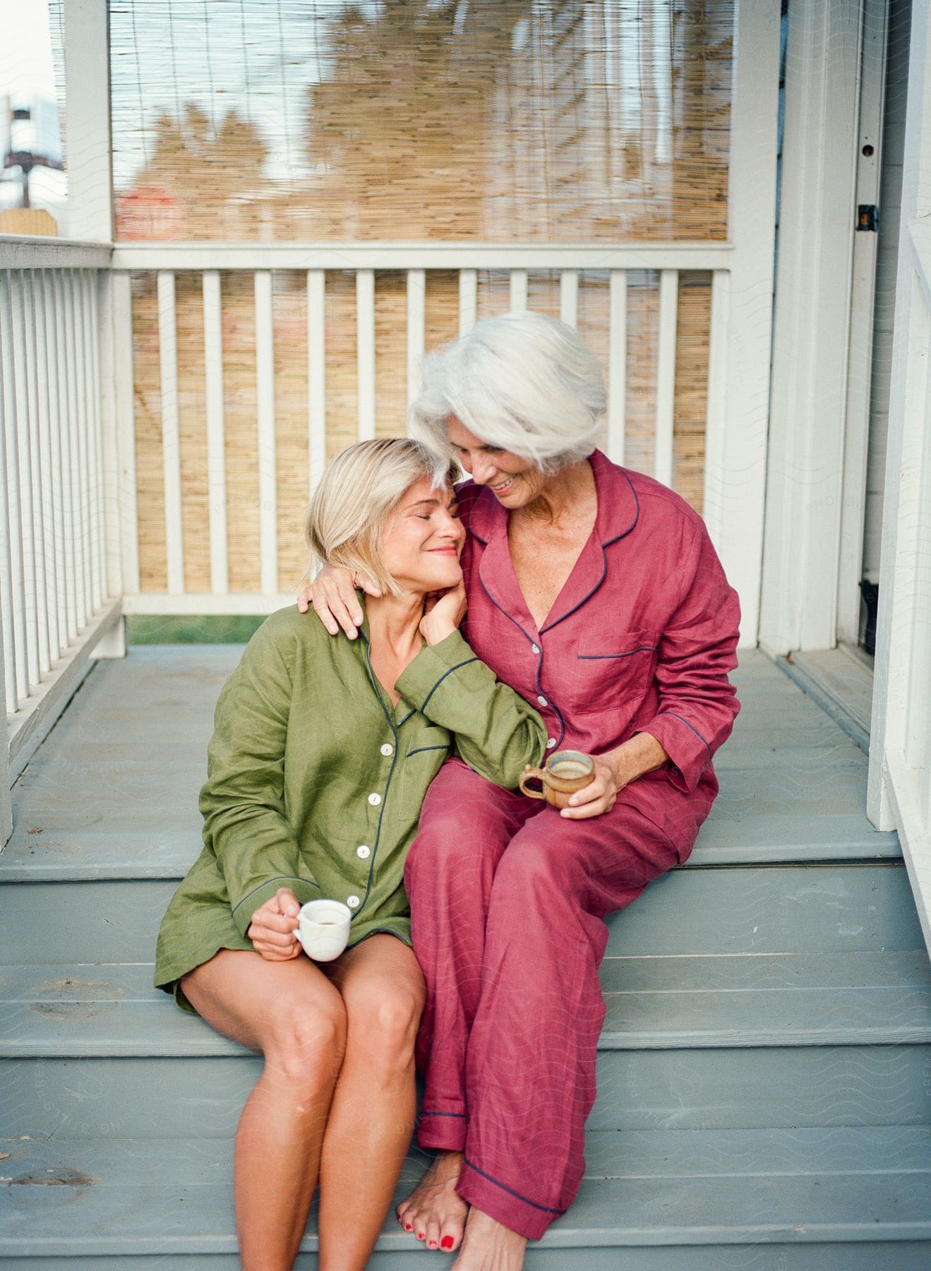 Woman sitting on the porch steps with her mother's arm around her as they hold coffee cups and smile