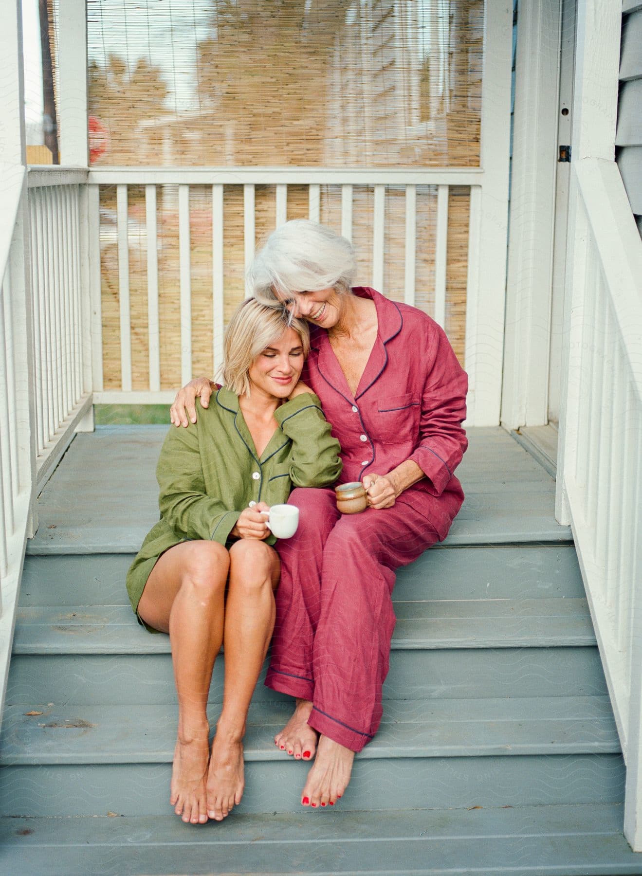 Mother and daughter in pajamas sitting together on a porch step while hugging each other affectionately and holding cups.