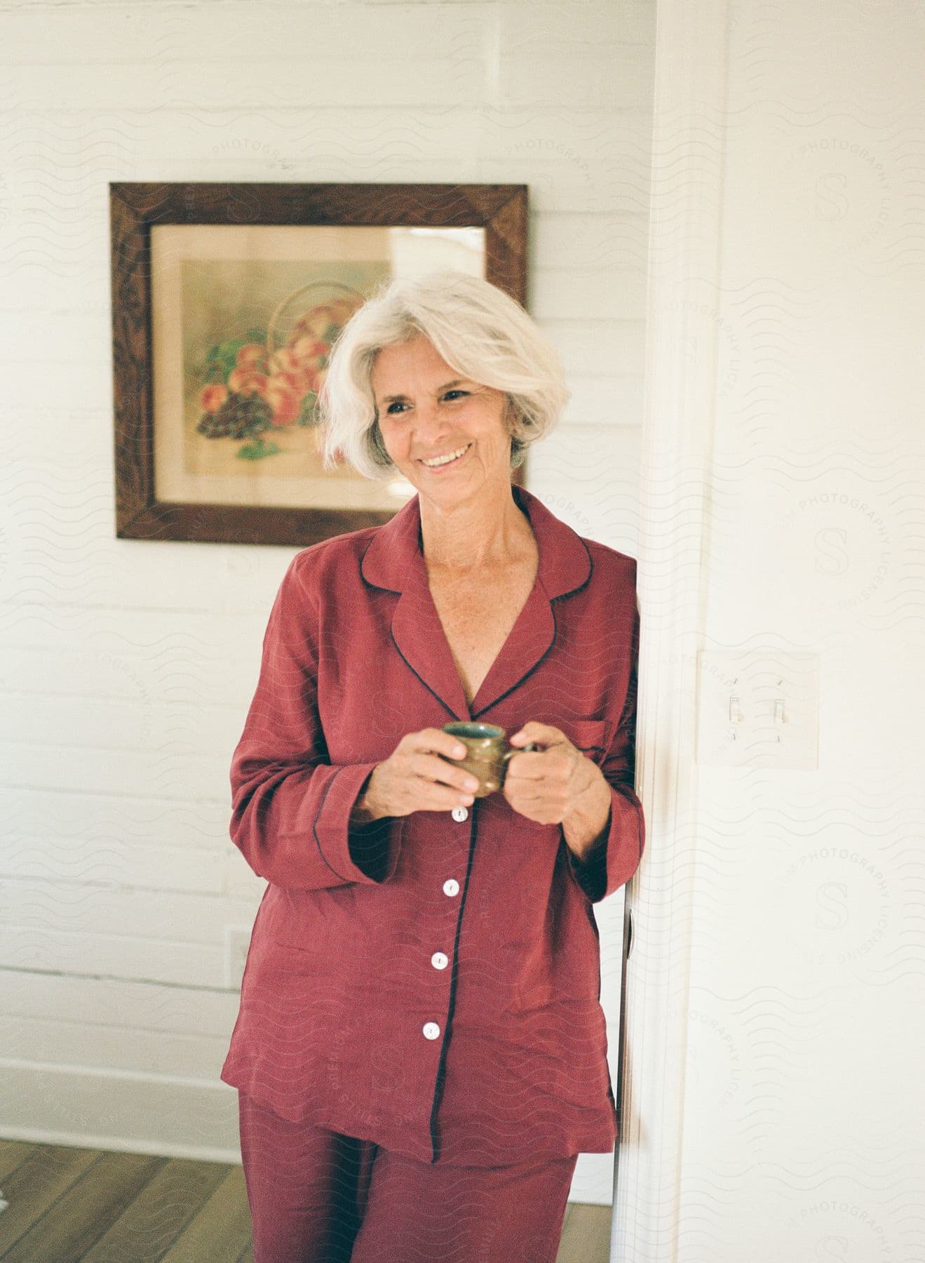 An elderly woman leans against the wall, holding a cup of coffee as she smiles.