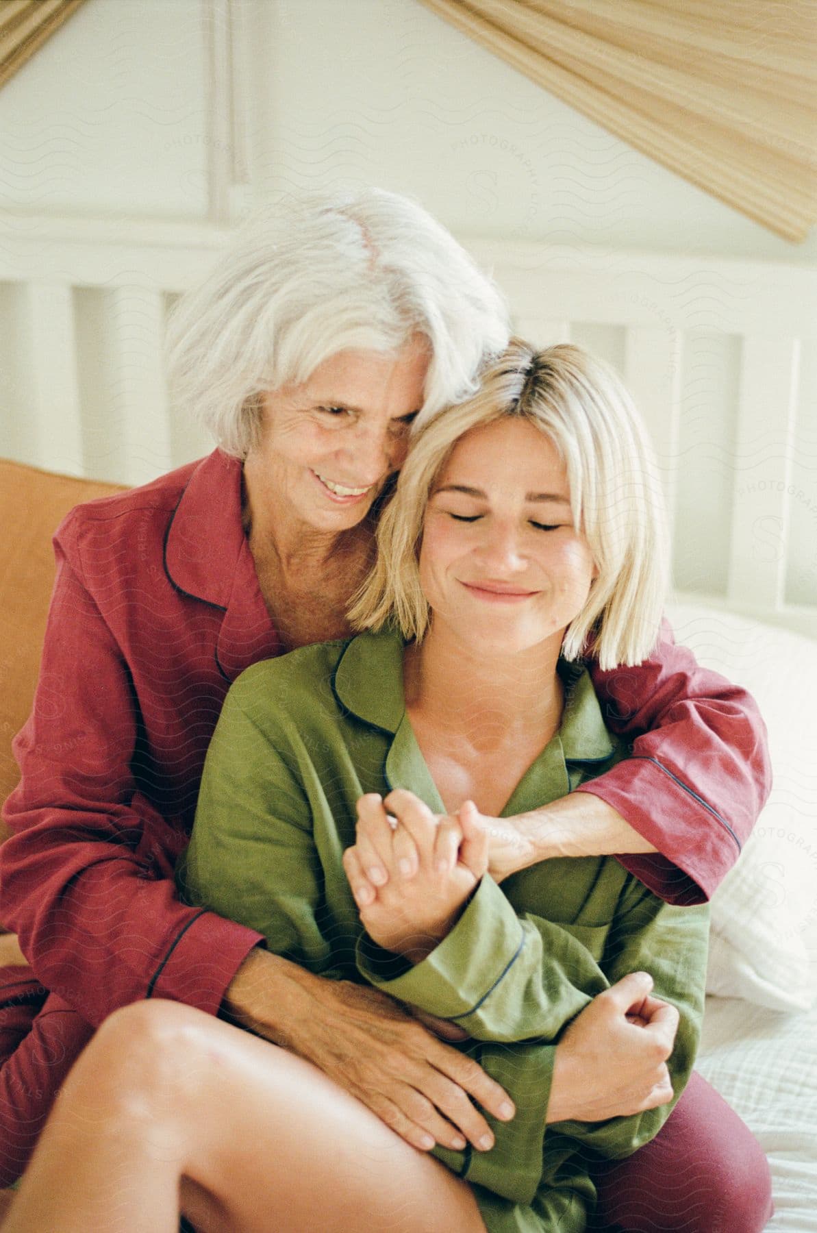 White haired mother smiles while sitting on bed and holding her grown daughter.